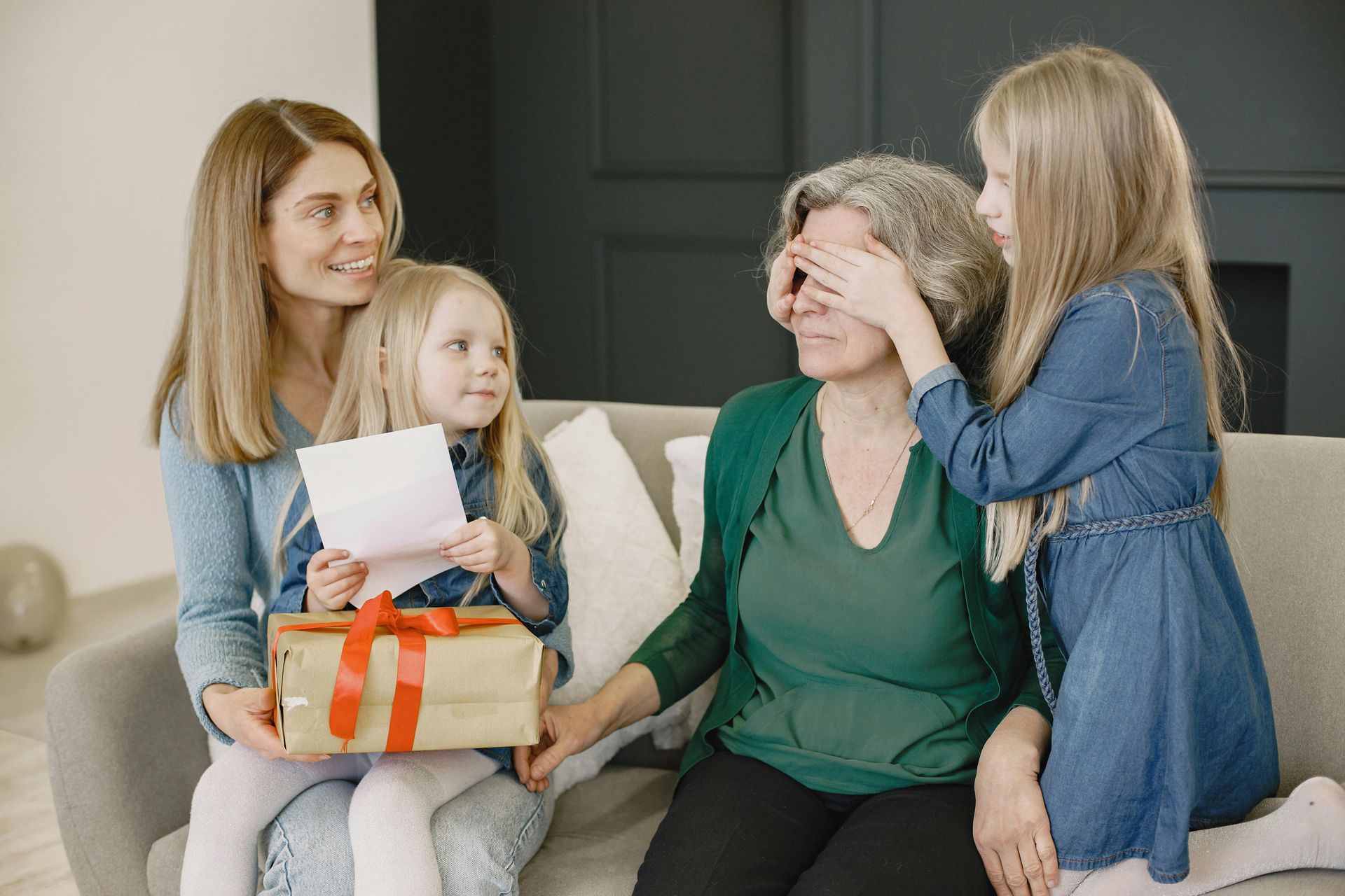 A child covers an adult's eyes while another child holds a gift, sitting together on a sofa with an adult family member.