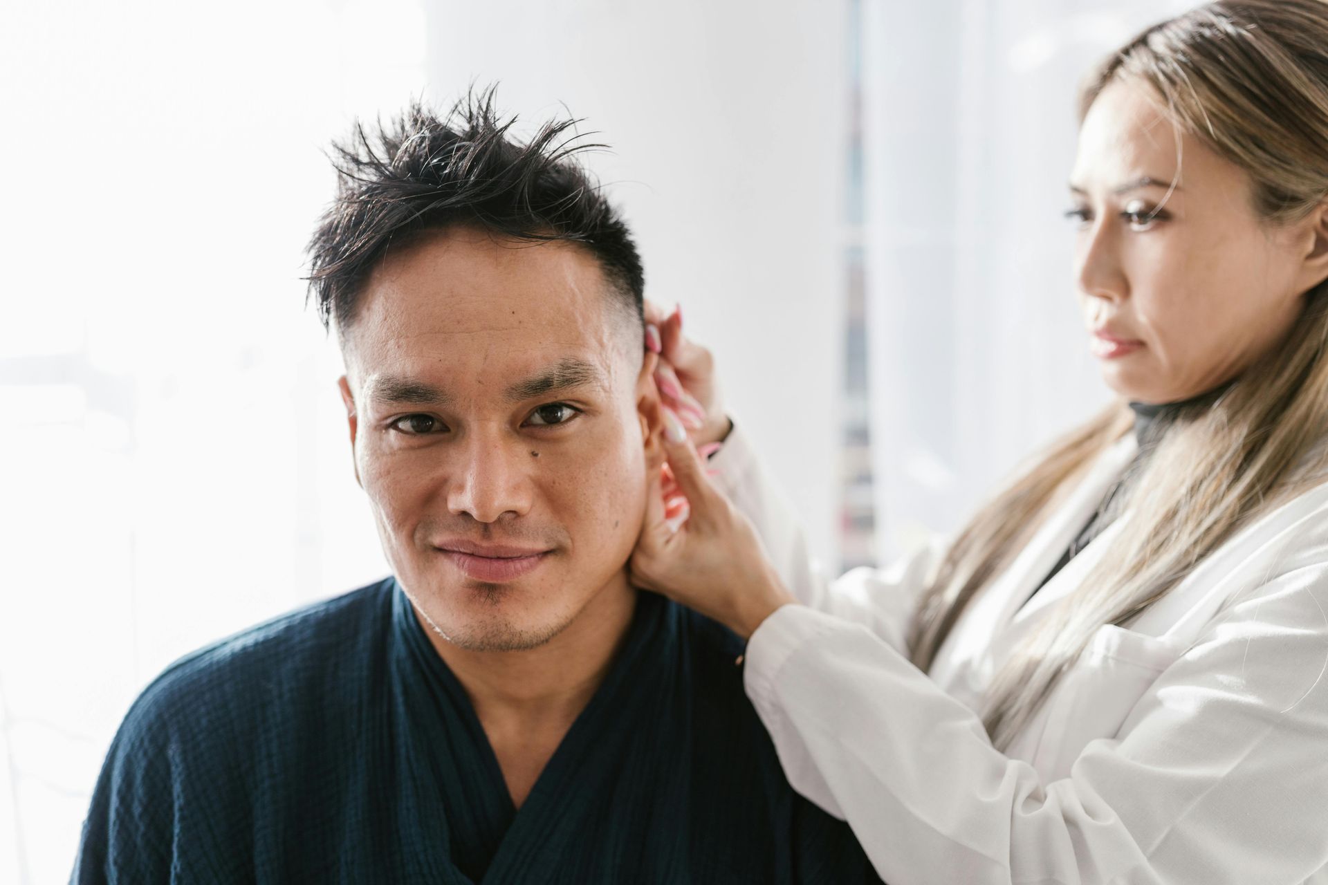 A person in a white lab coat examines the ear of a man wearing a dark robe in a bright, clinical setting.