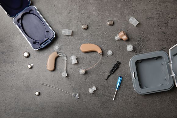 Two beige behind-the-ear hearing aids, accessories, batteries, and a small screwdriver on a gray surface.
