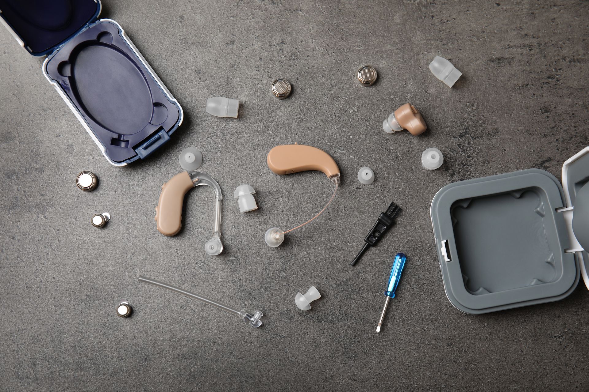Two beige hearing aids, ear domes, batteries, cleaning tools, and two small cases arranged on a gray surface.