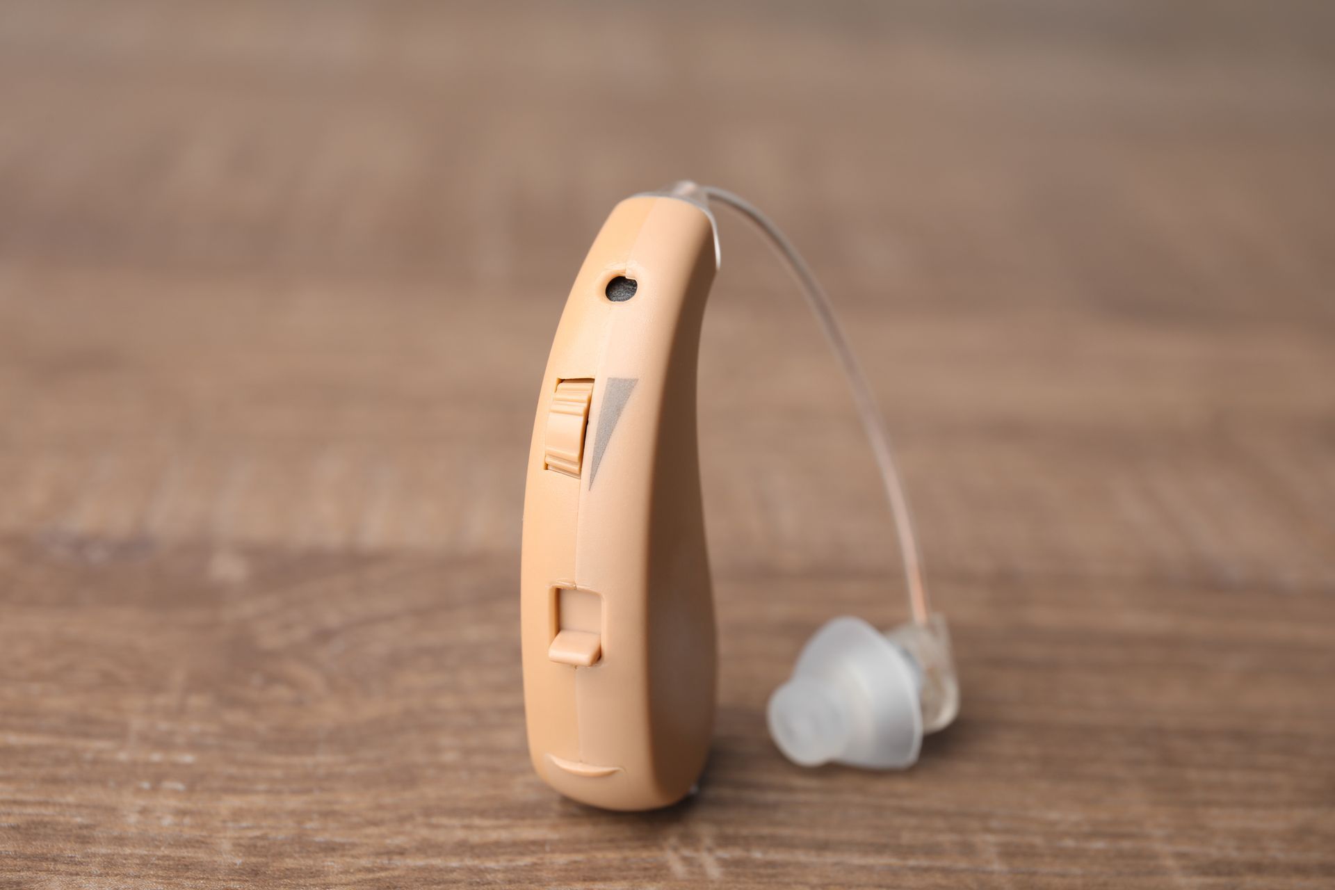 A beige behind-the-ear hearing aid with a clear ear tip, resting on a wooden surface.