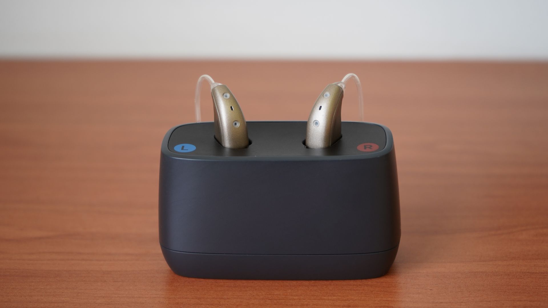 Two beige hearing aids sitting in a black charging station on a wooden surface.