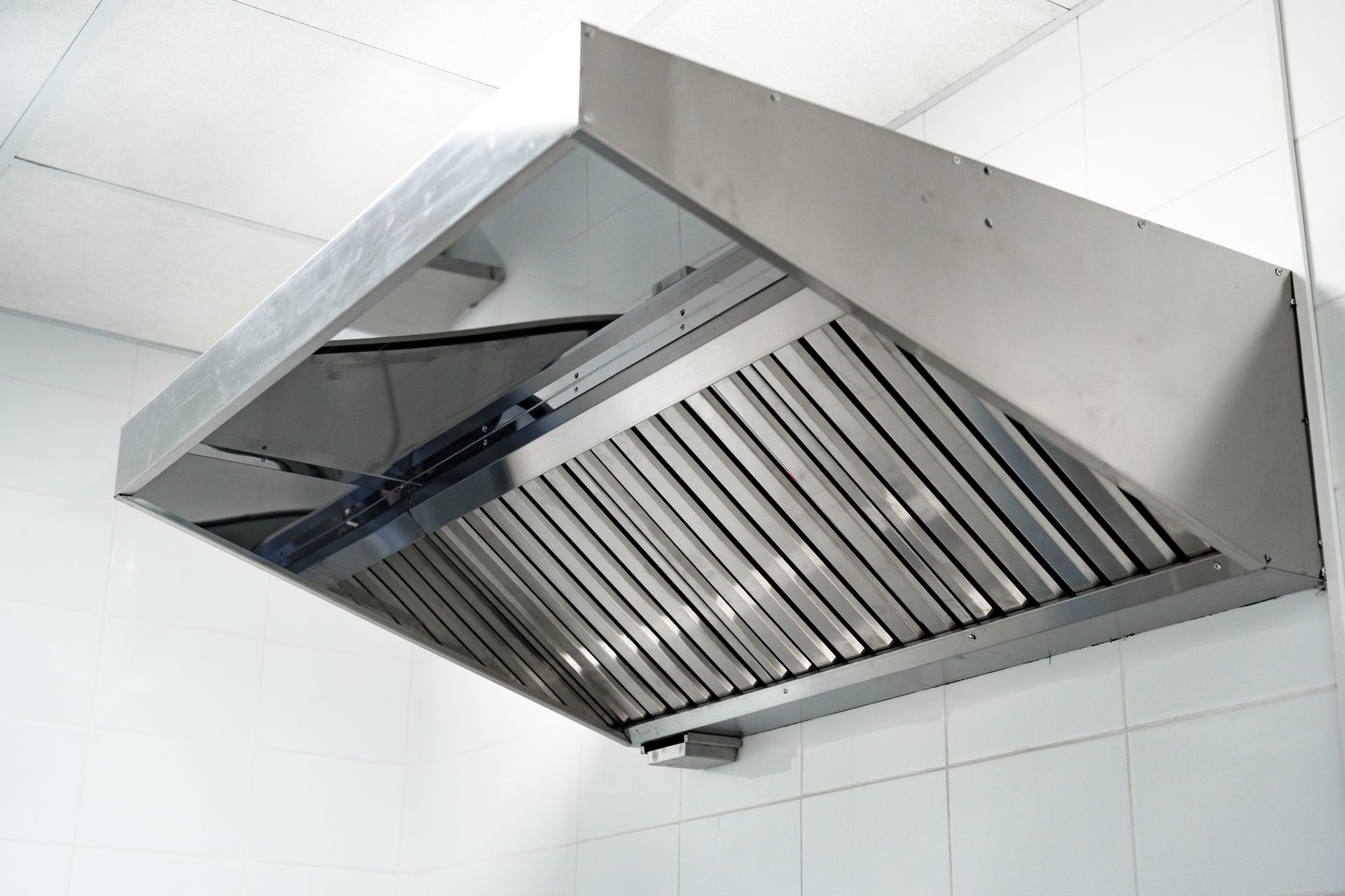 Stainless steel commercial kitchen exhaust hood mounted on a white tiled wall.
