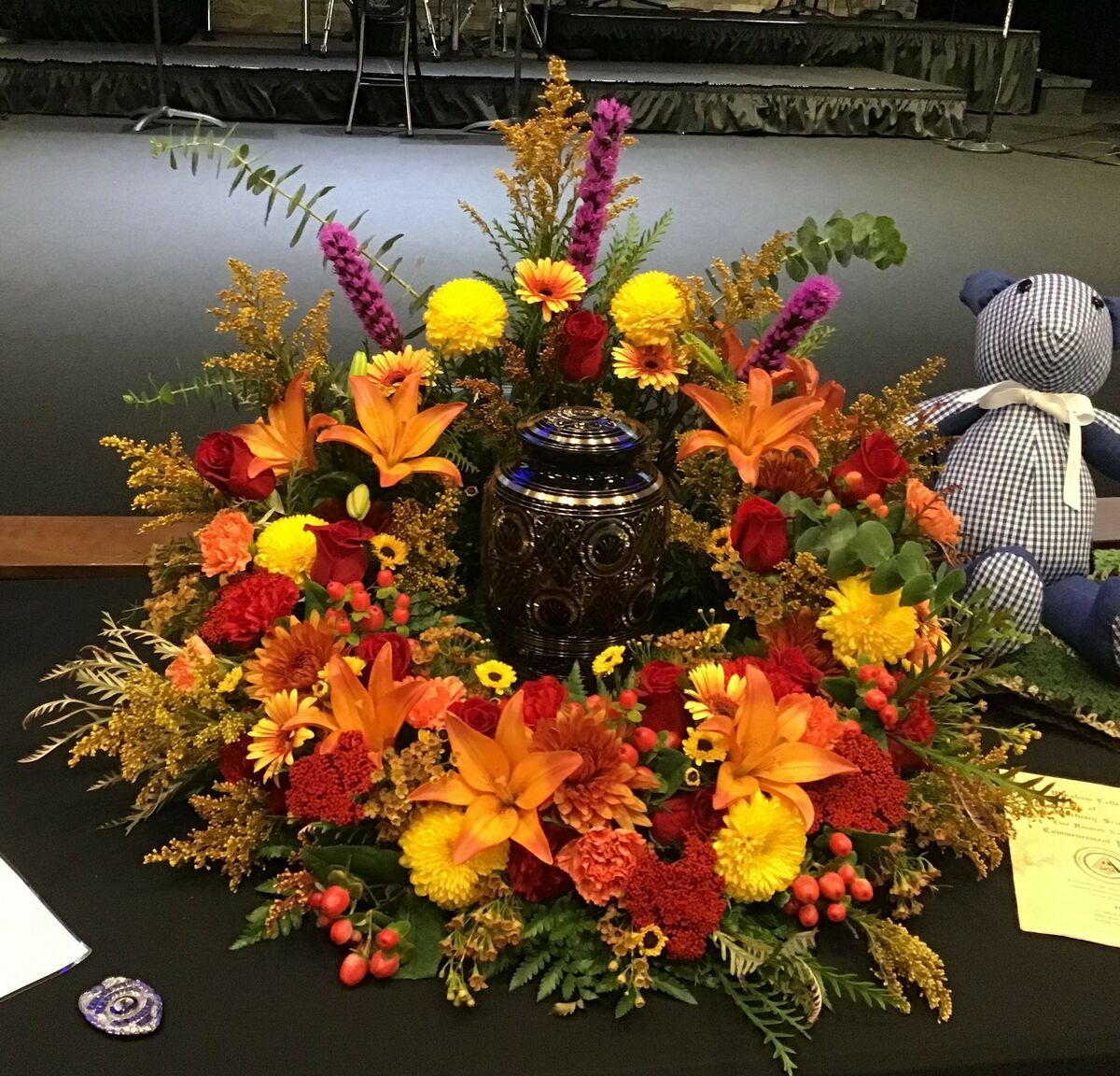 A teddy bear sits on a table next to a wreath of flowers