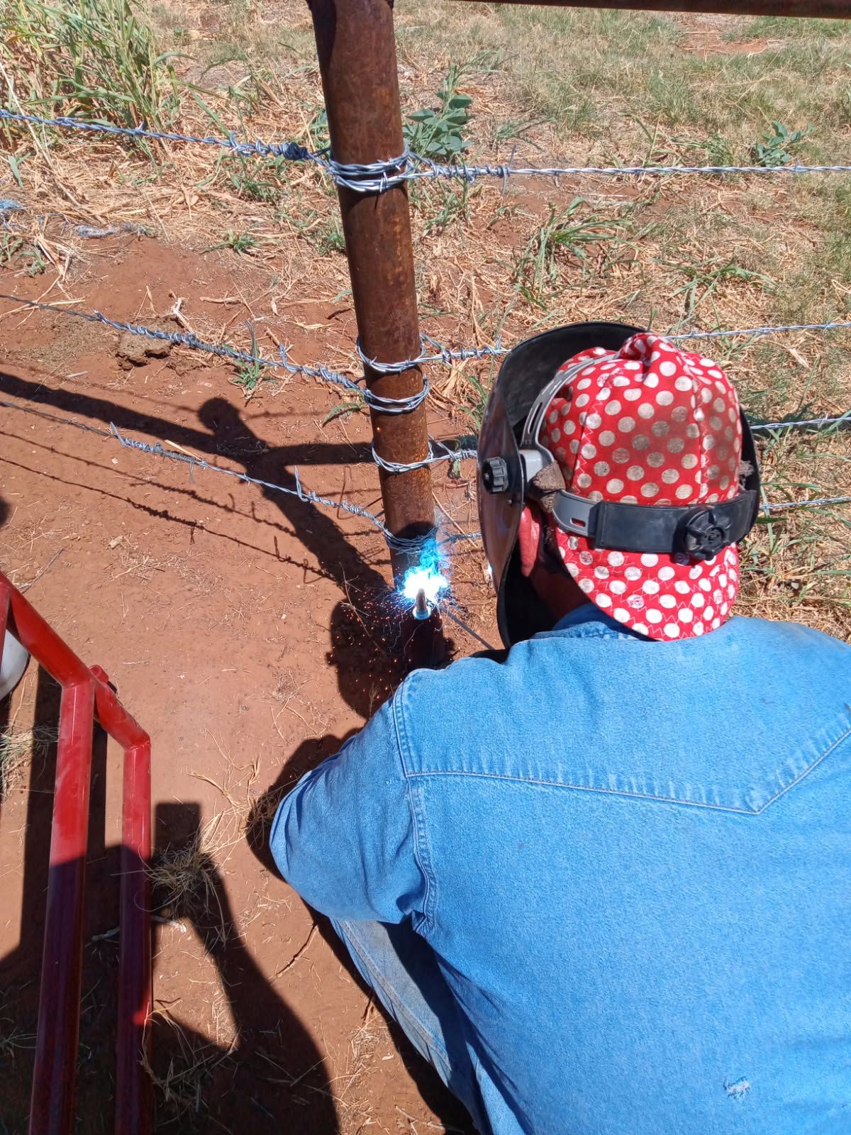 Person welding metal fence post outdoors, wearing a hat and safety gear.