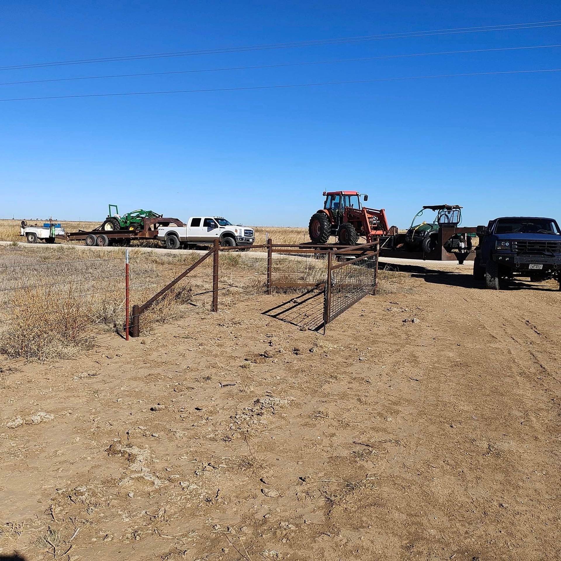 Dirt road with gate, vehicles and farm equipment under a blue sky.