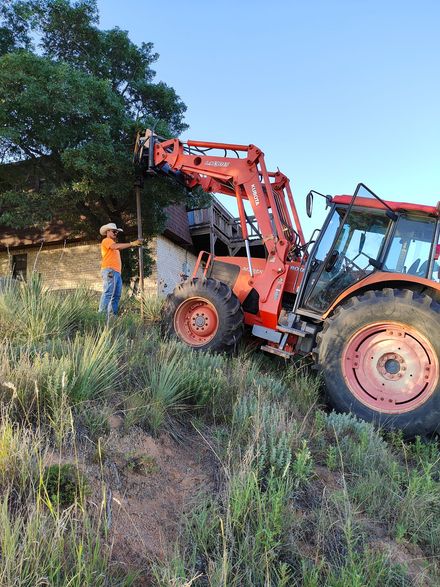 Man in orange shirt uses tractor to clear vegetation from a brick building.
