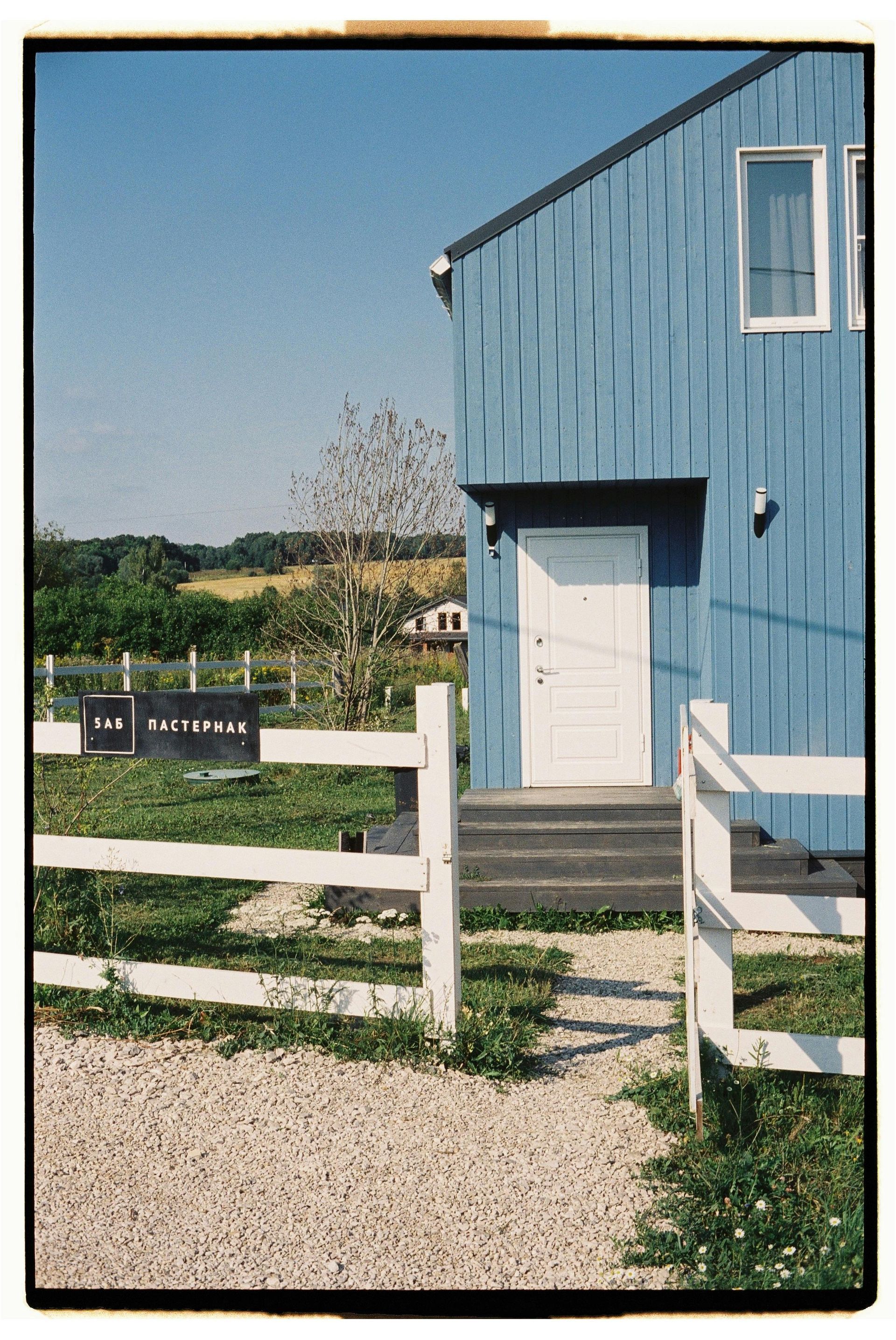 Blue house with white door and fence, gravel path, sunny field in background.