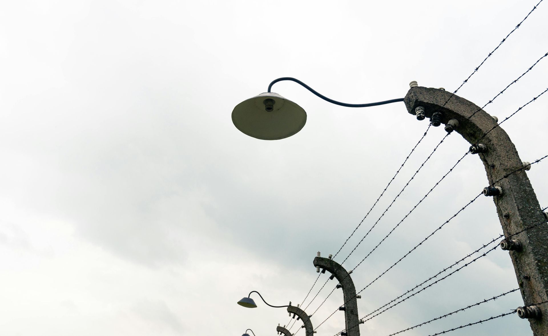 Barbed wire fence with a lamppost against a cloudy sky, likely at a historical site.