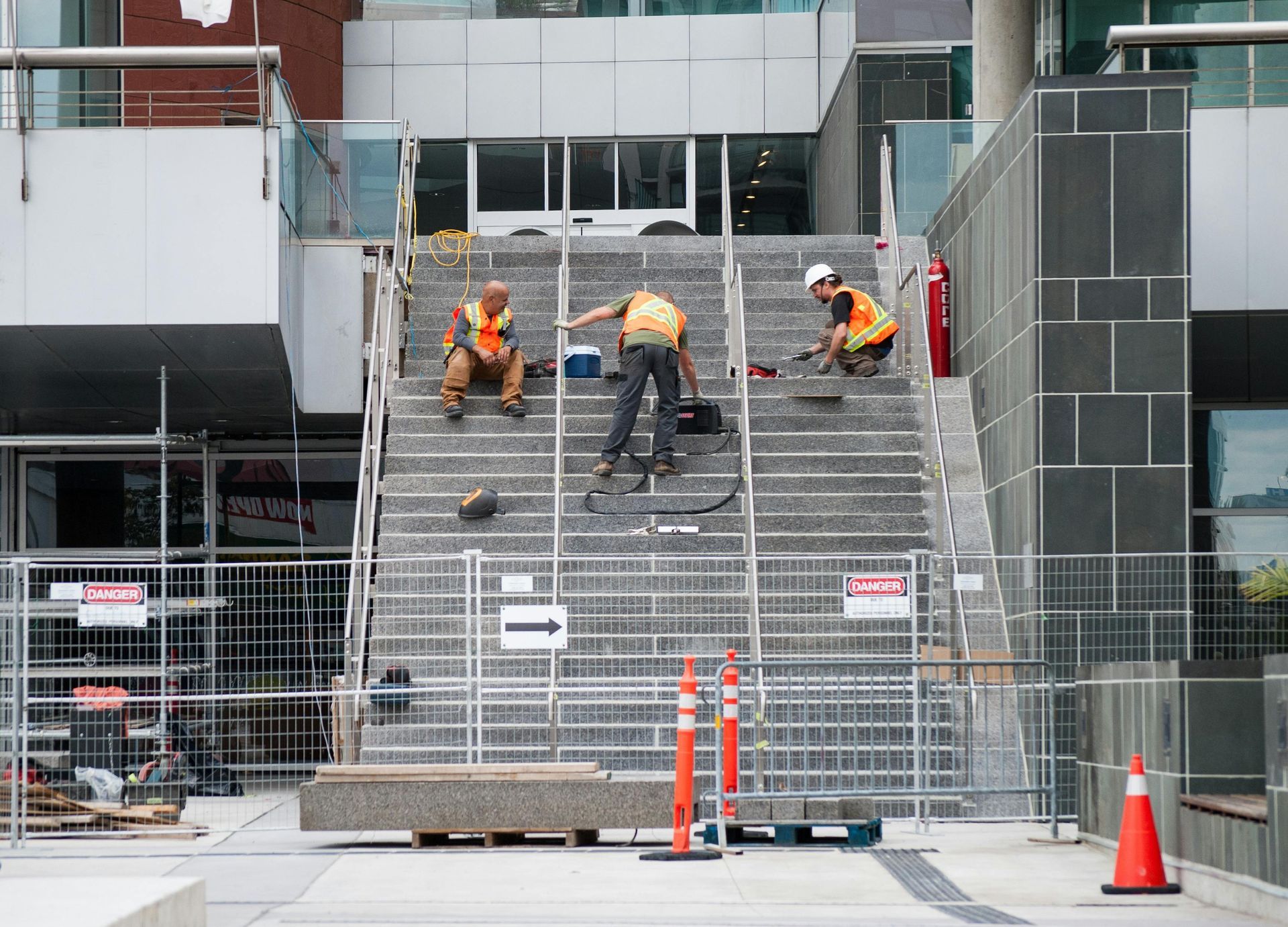 Construction workers on steps, wearing orange vests and hard hats, working on a building exterior.