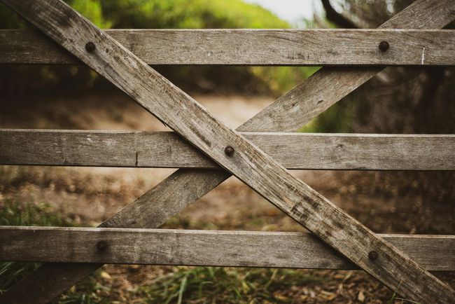 Wooden gate, weathered and closed, blocks a dirt path in a natural outdoor setting.