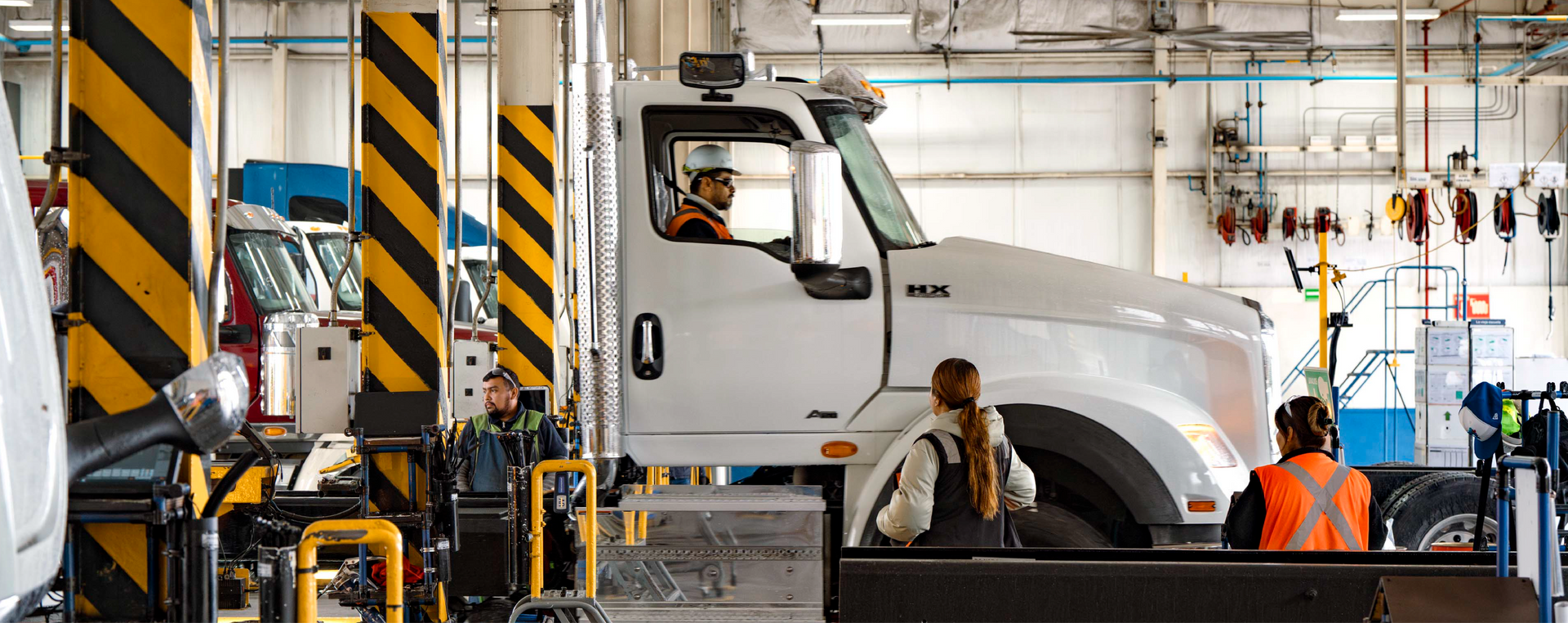 White truck in a service bay, a person in driver's seat. Two workers wearing orange vests and safety glasses are nearby.