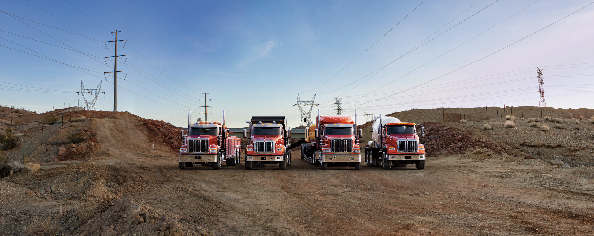 Four red semi-trucks parked on a dirt road in front of hills, with power lines and blue sky.