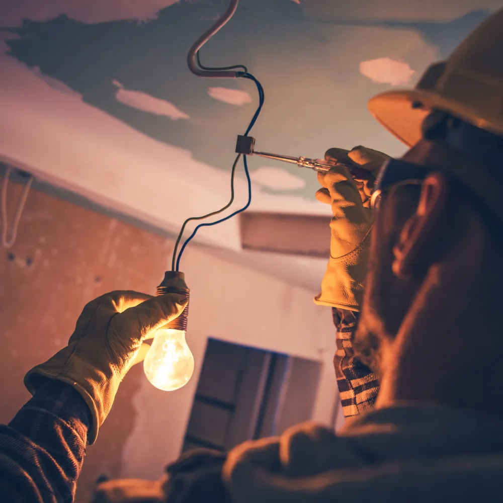 Electrician installing a light bulb, using a screwdriver, wearing gloves and a hard hat.