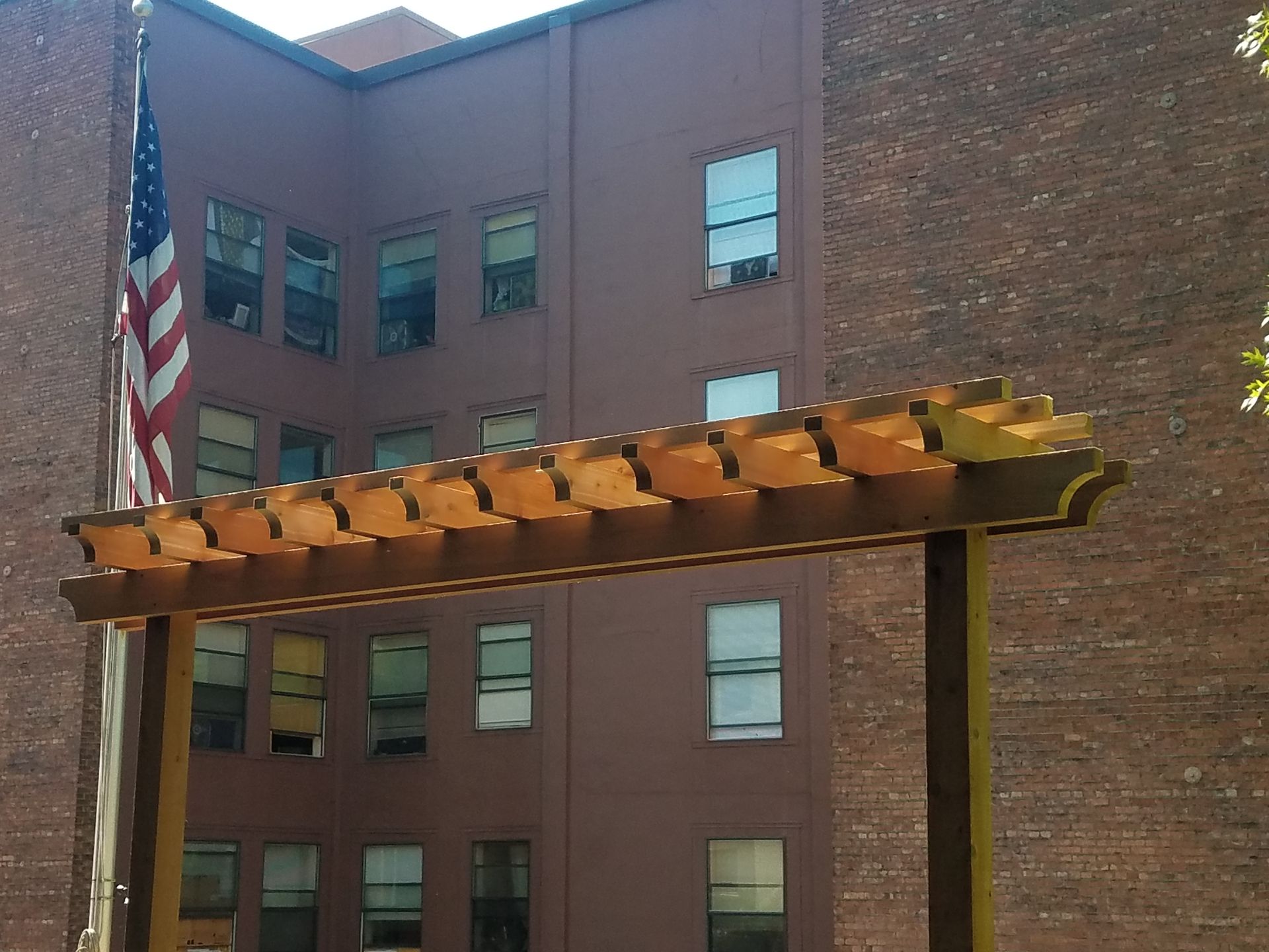 Wooden pergola structure with a brown building and an American flag in the background.