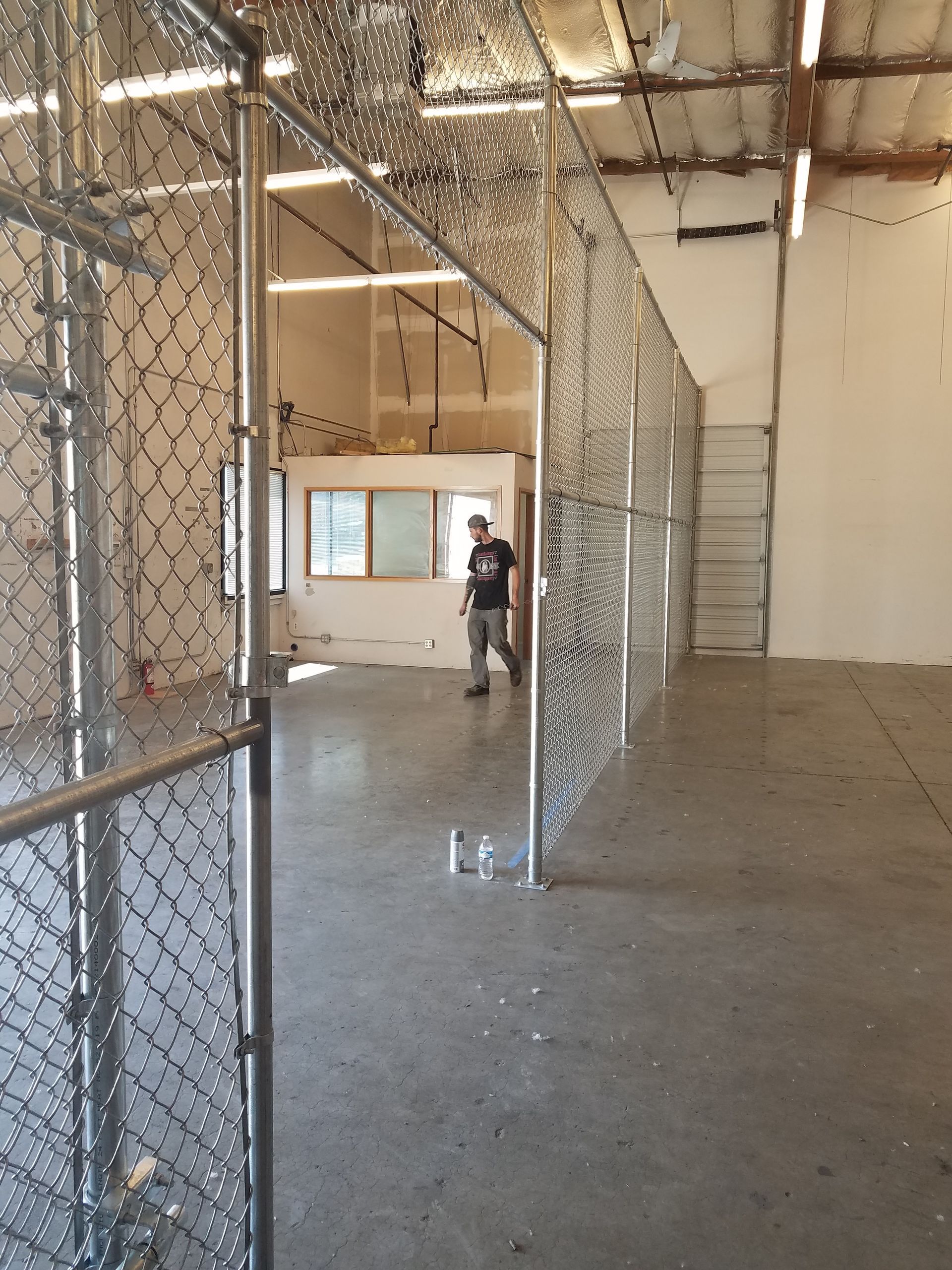 Interior view of a building with chain-link fences. A person stands near a window. Concrete floor, walls, and metal structures.