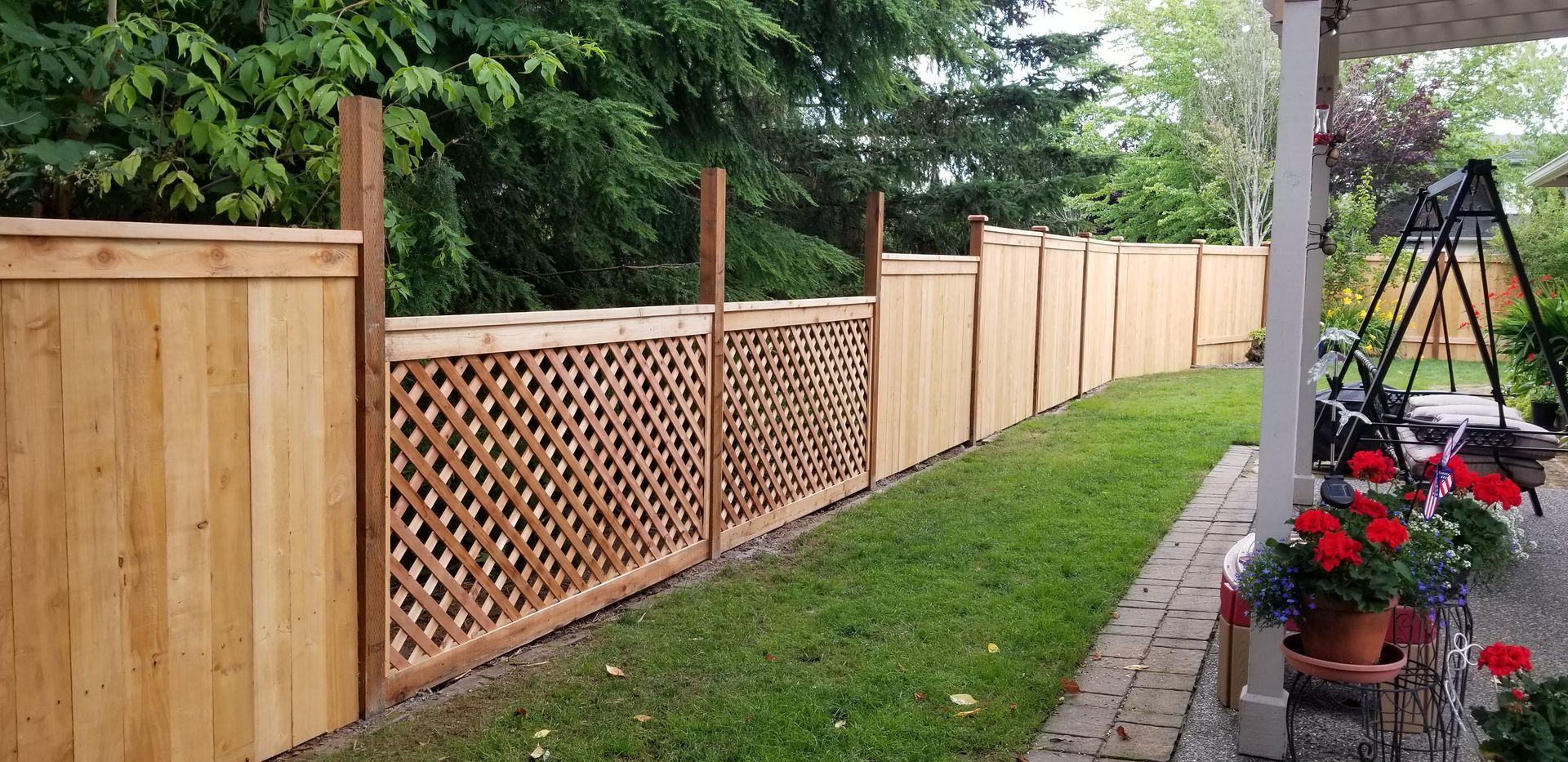 A wooden fence with lattice and solid panels, bordering a green lawn. Flowers in pots and a patio.