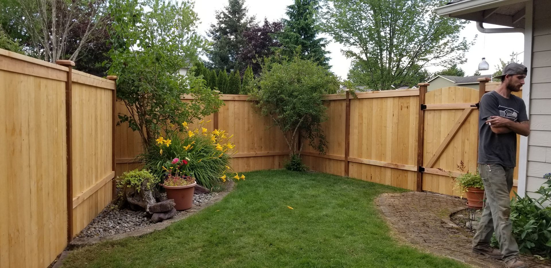 Wooden fence encloses a small backyard with green grass, plants, and a person standing near a building.