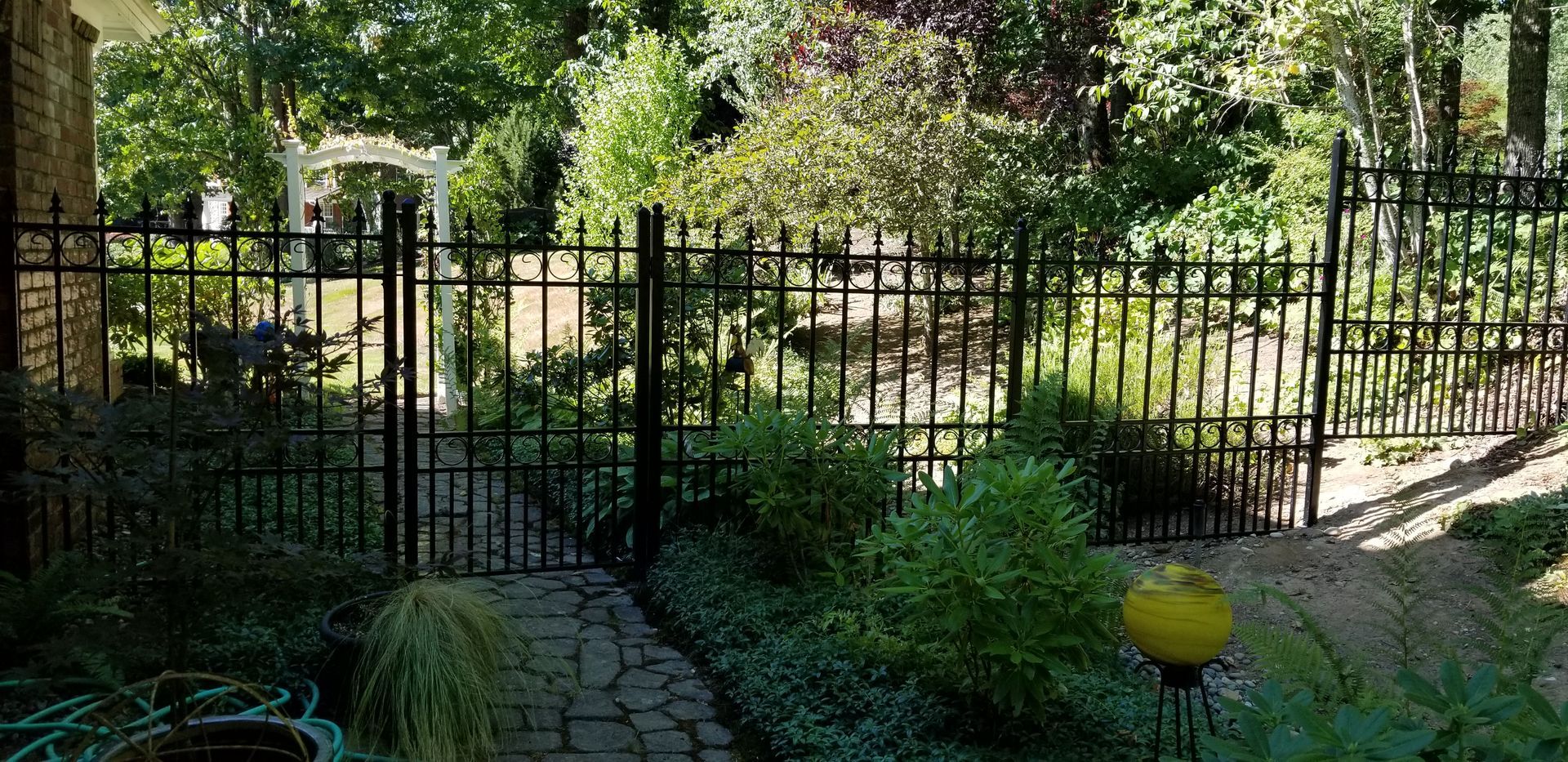 Black wrought iron fence with gate, brick pathway, and lush greenery in a garden setting.