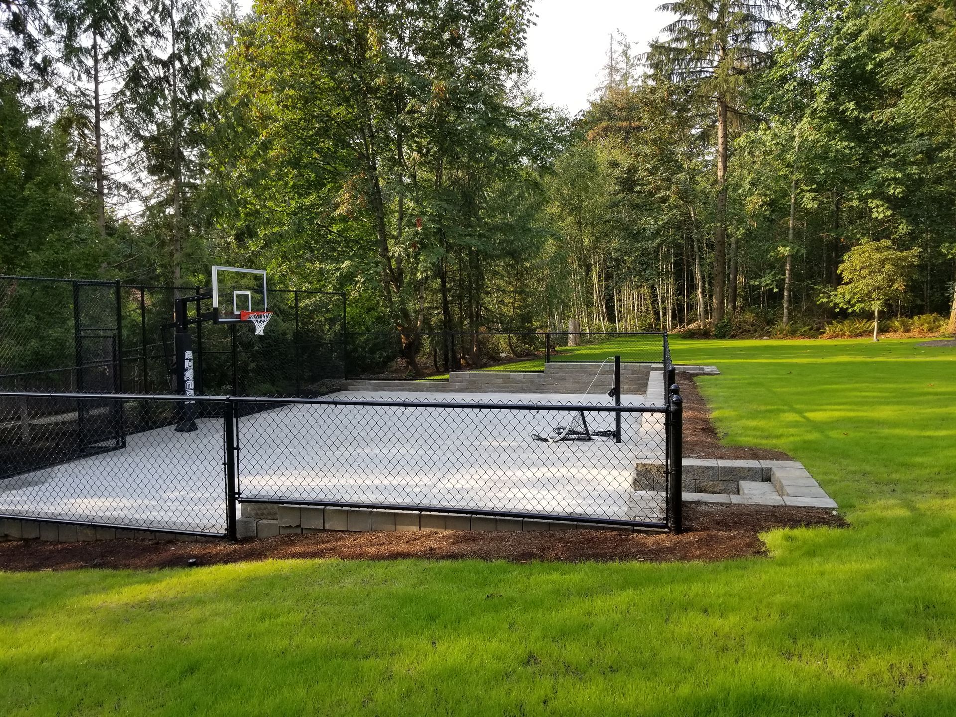 Outdoor basketball court enclosed by black fence, surrounded by grass and trees.