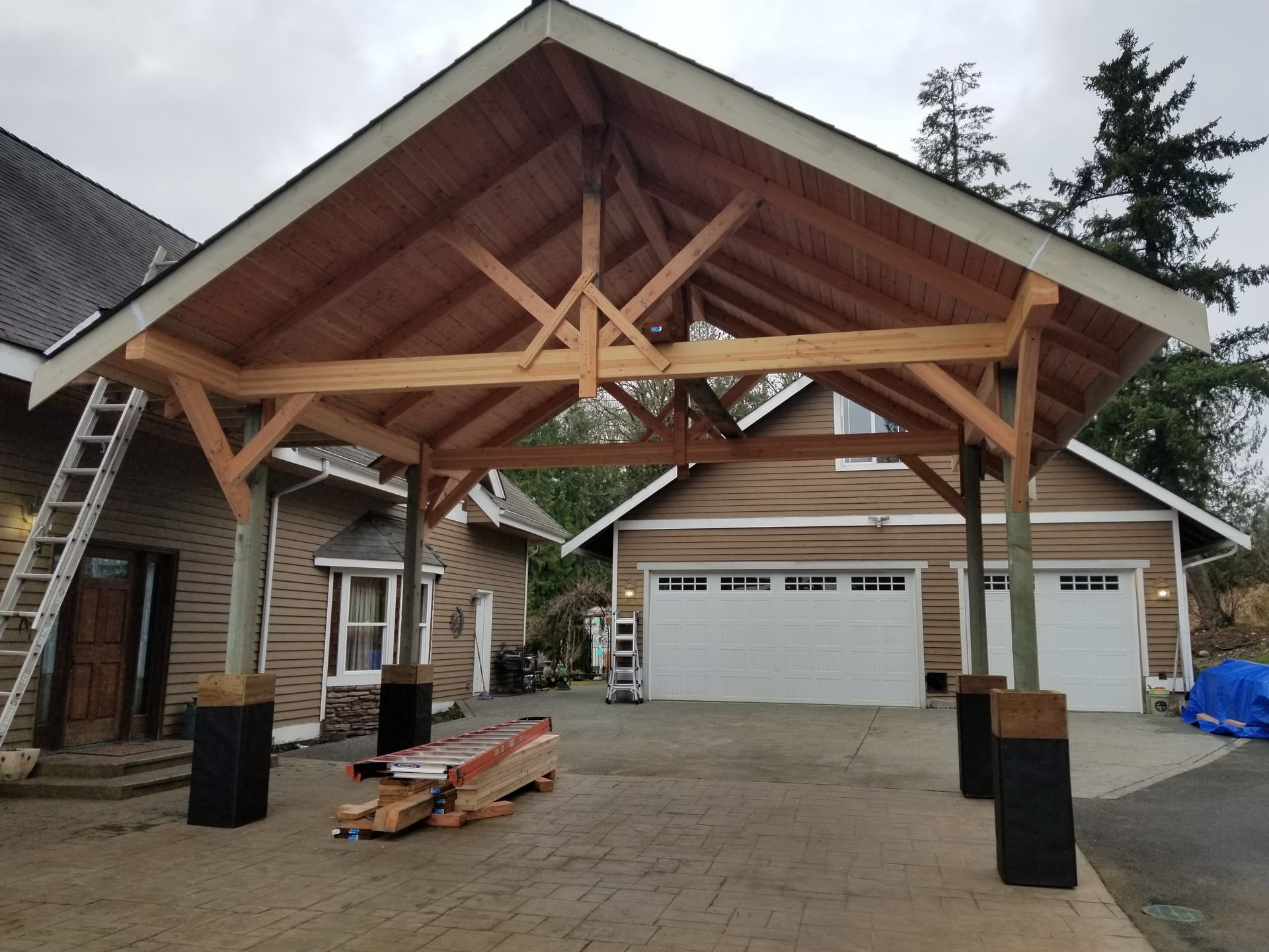 Wooden carport in driveway connecting to a house and garage with white doors.