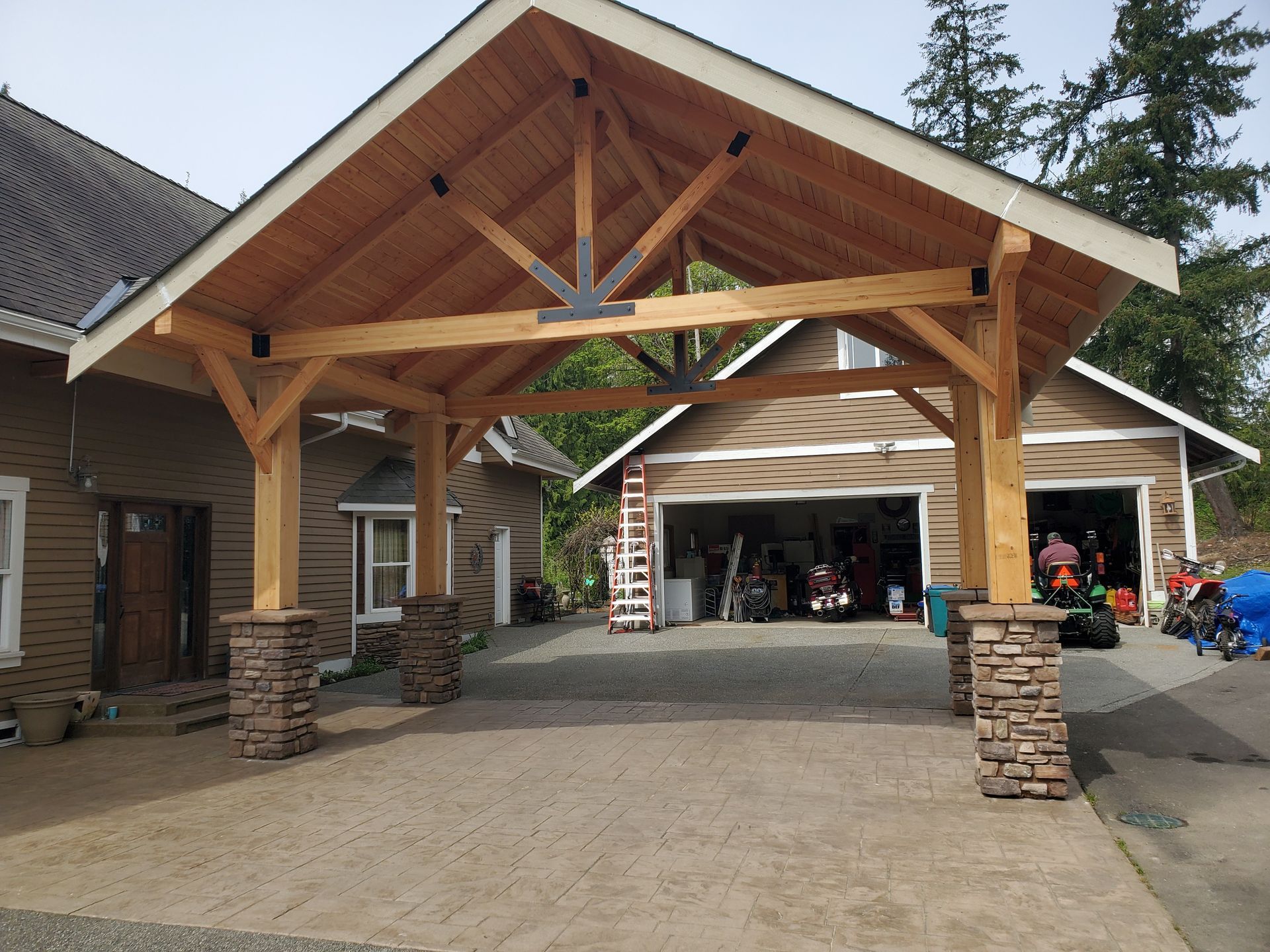 Wooden carport with brown roof, supported by stone-covered posts, over a concrete driveway. A garage is visible in the background.