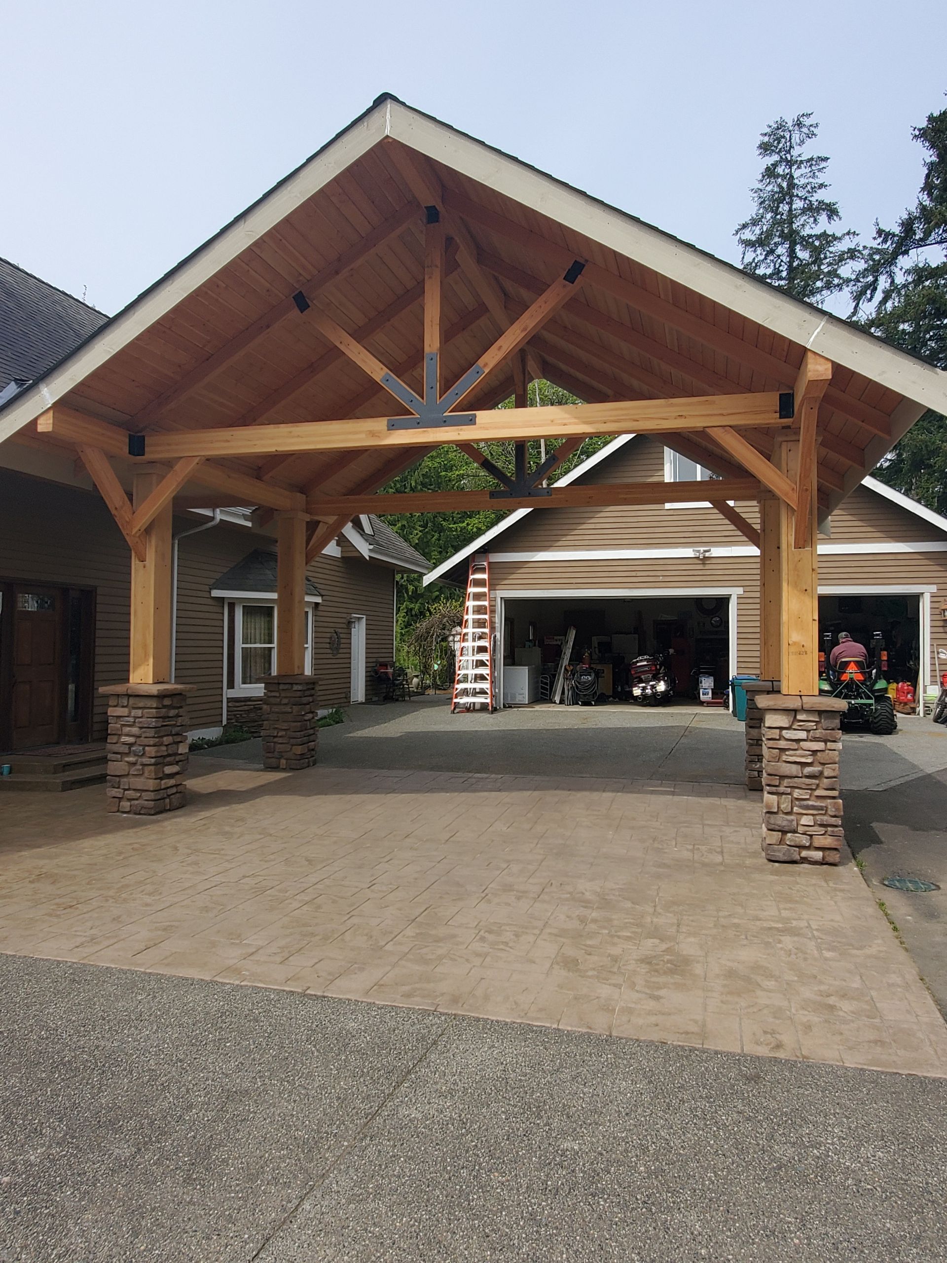 Carport over driveway, connected to house and garage. Wooden frame with stone-covered columns.