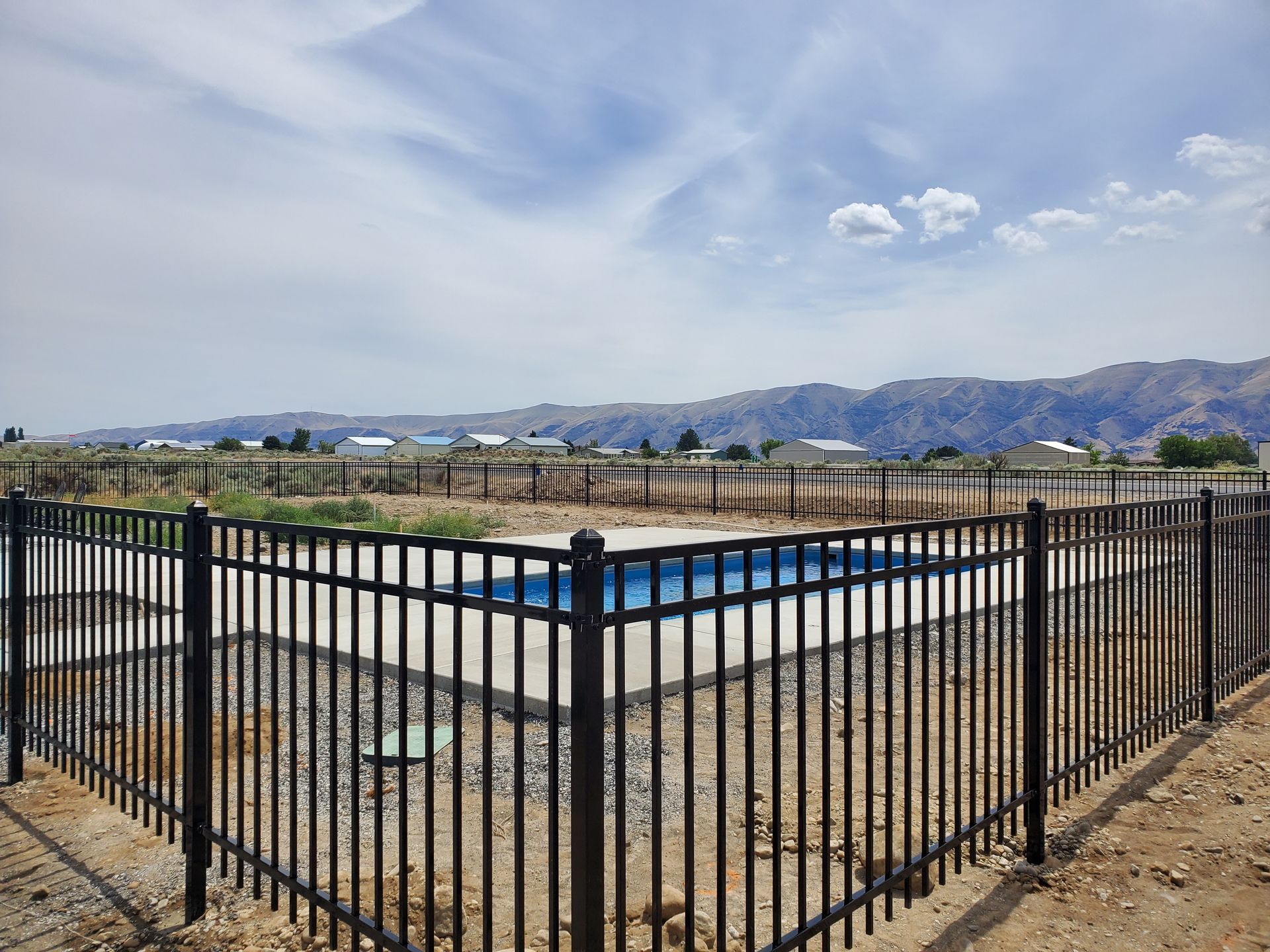 Black fenced-in rectangular pool in a desert setting with mountains in the background under a partly cloudy sky.