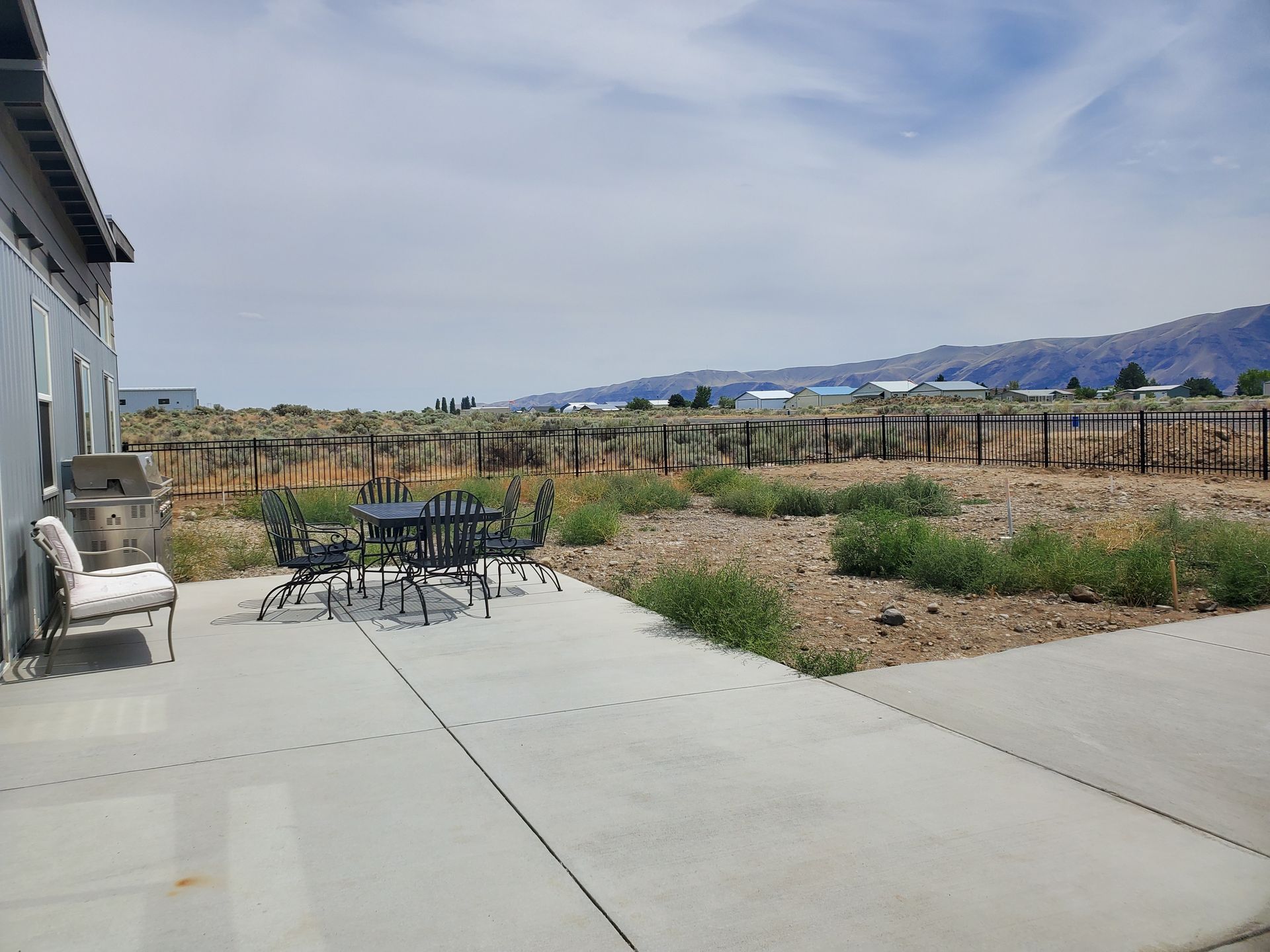 Concrete patio with outdoor table and chairs, overgrown dirt yard, distant buildings and mountains.