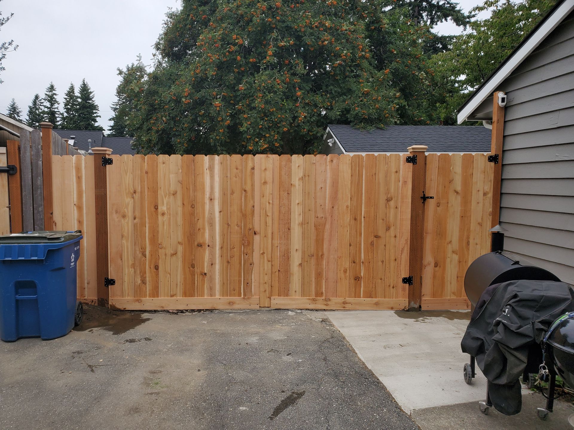 Wooden fence with gate, blue trash bin, and barbecue grill on a concrete surface.