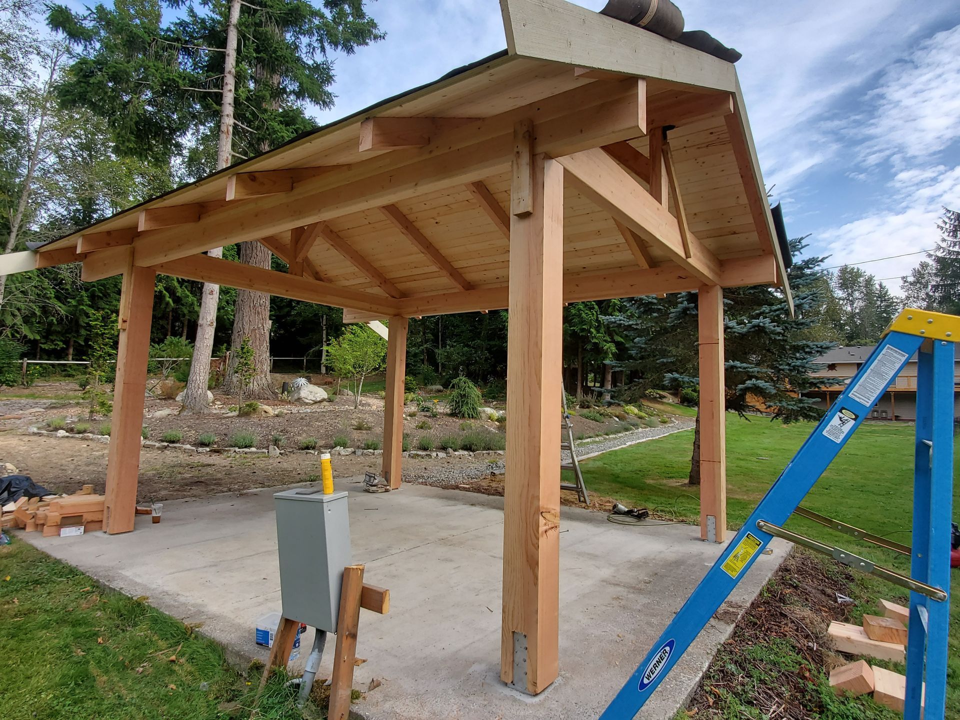 Wooden gazebo under construction on a concrete pad, surrounded by grass and trees.
