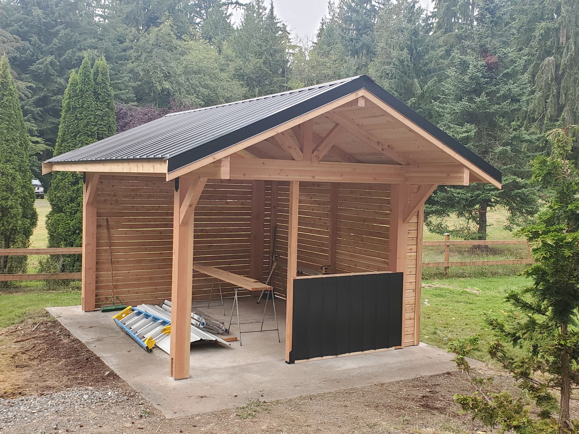 Wooden outdoor shelter with black roof, built on a concrete slab, set in a wooded area.