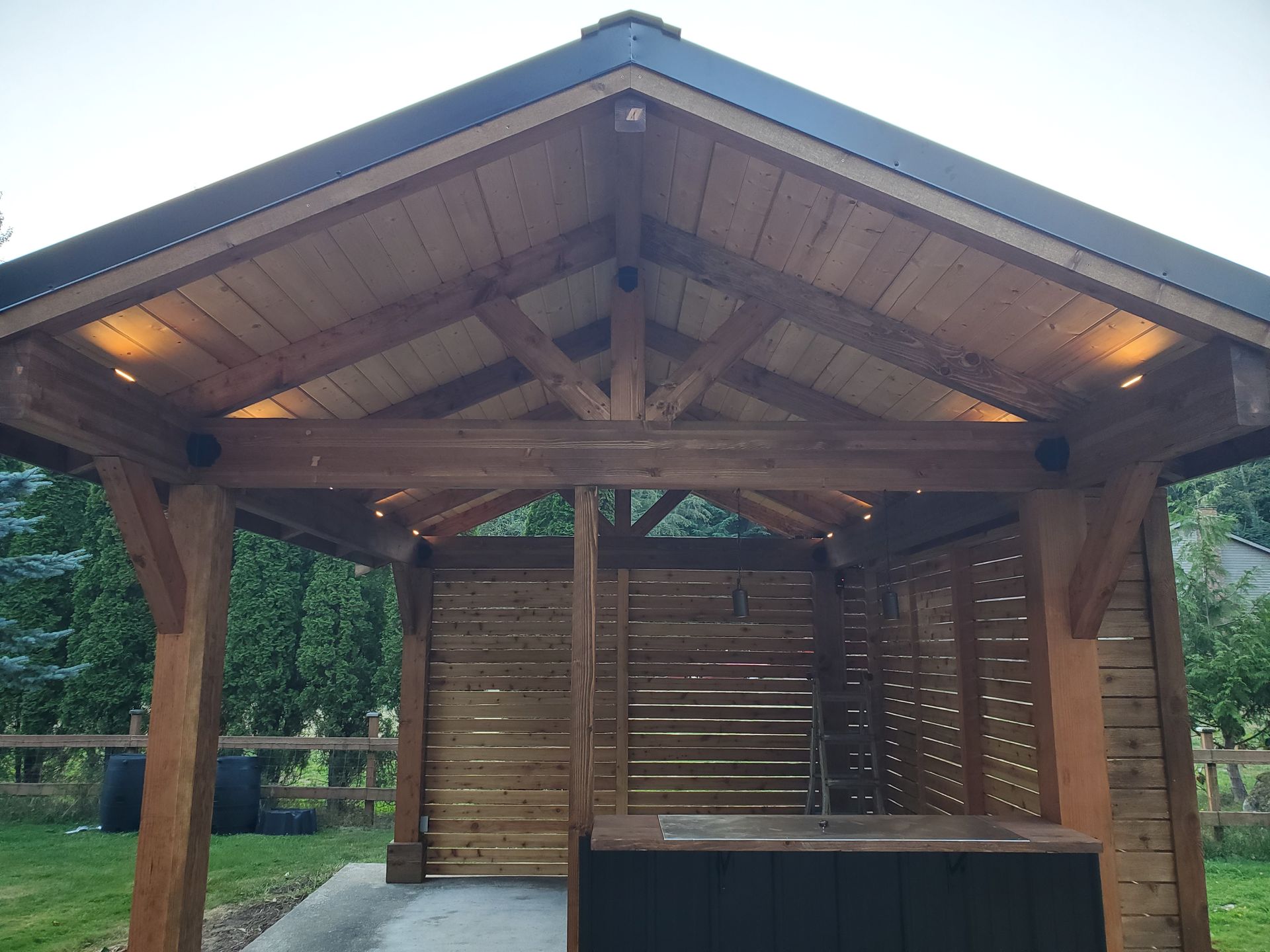 Wooden gazebo with a dark roof and built-in lighting, with a slatted wooden back wall, and concrete flooring.