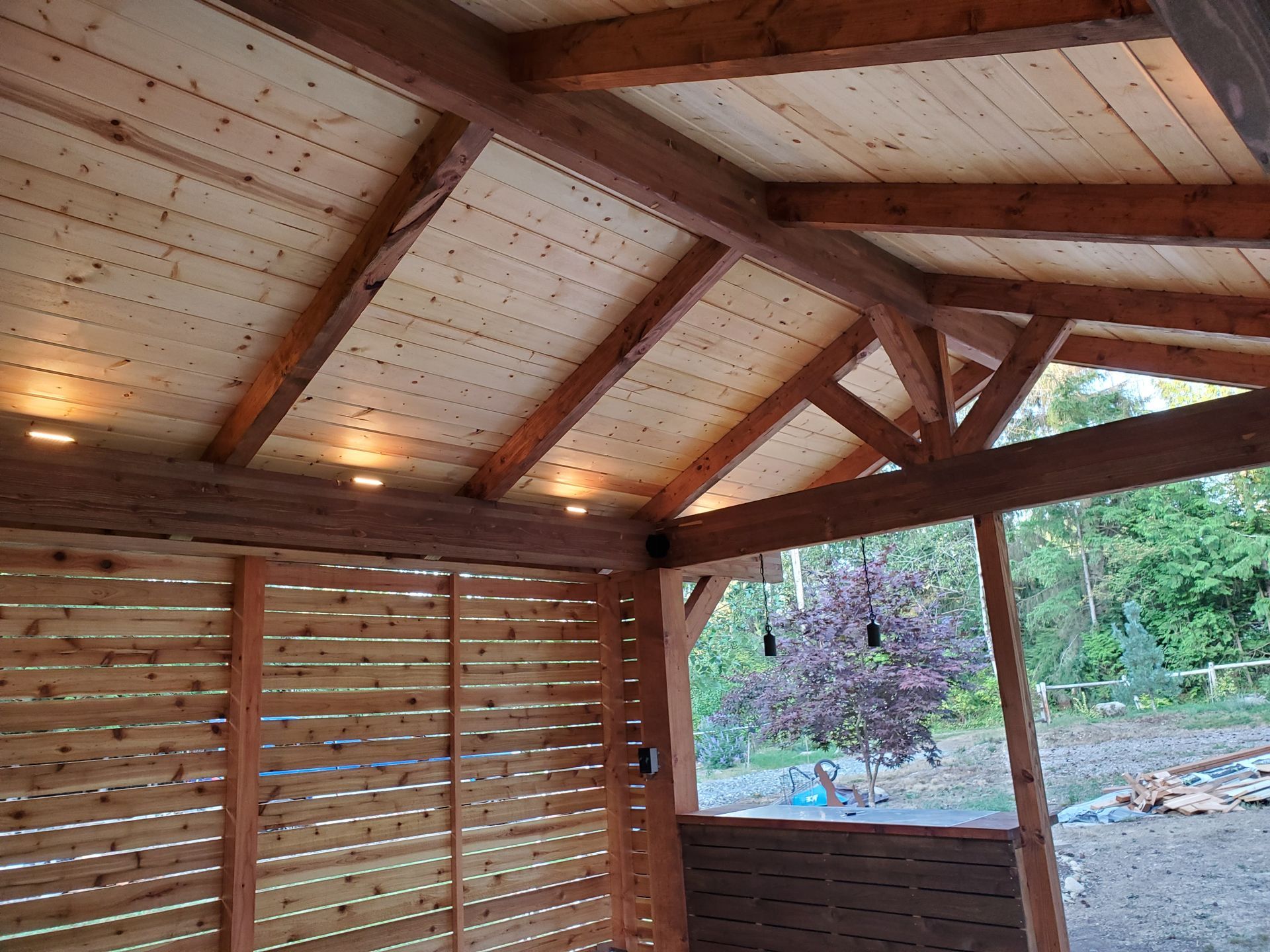 Wooden outdoor structure with slatted walls and ceiling, featuring built-in lights and a view of trees.