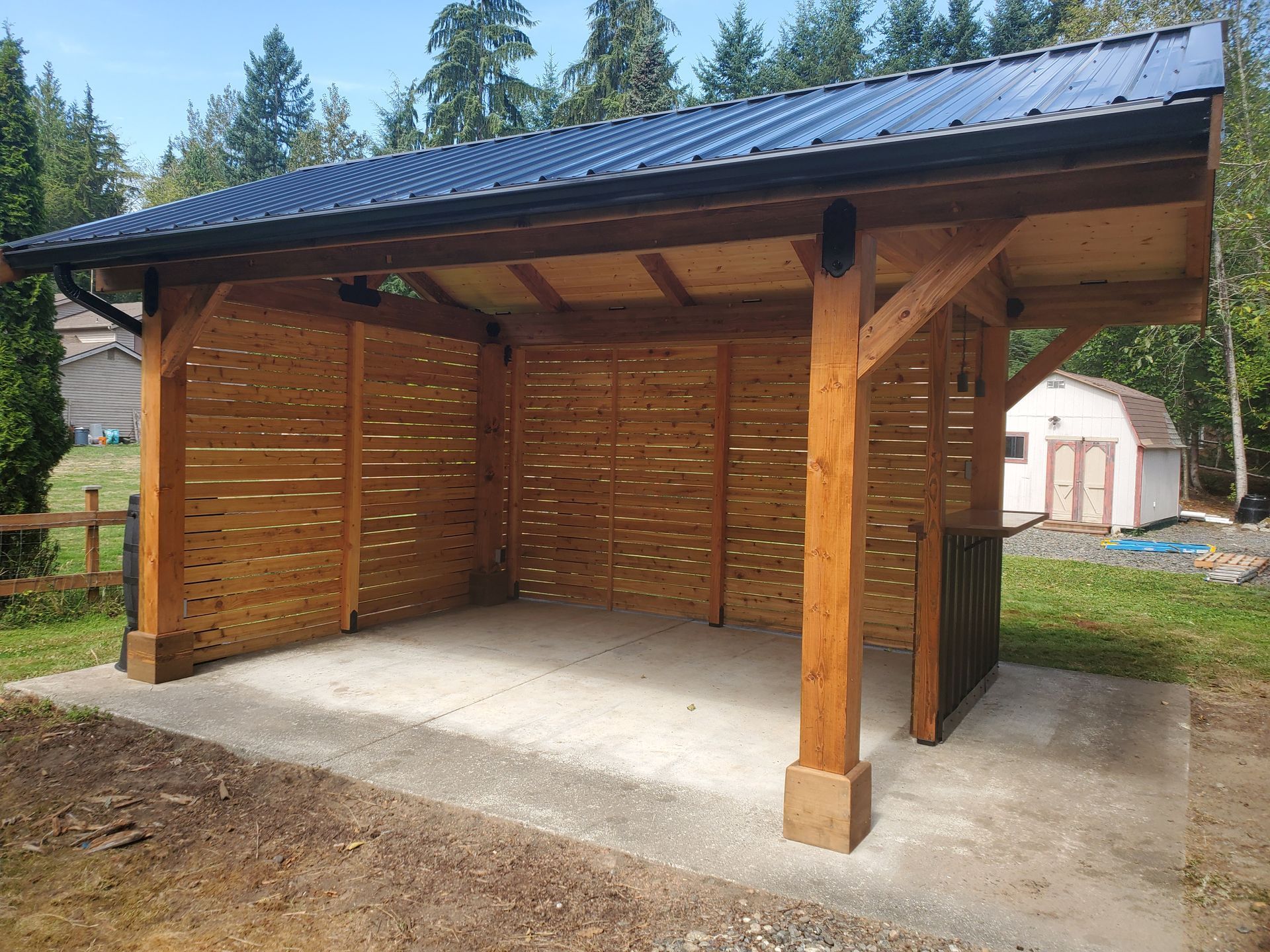Wooden carport with a black metal roof, concrete floor, and slatted walls. Backyard setting.