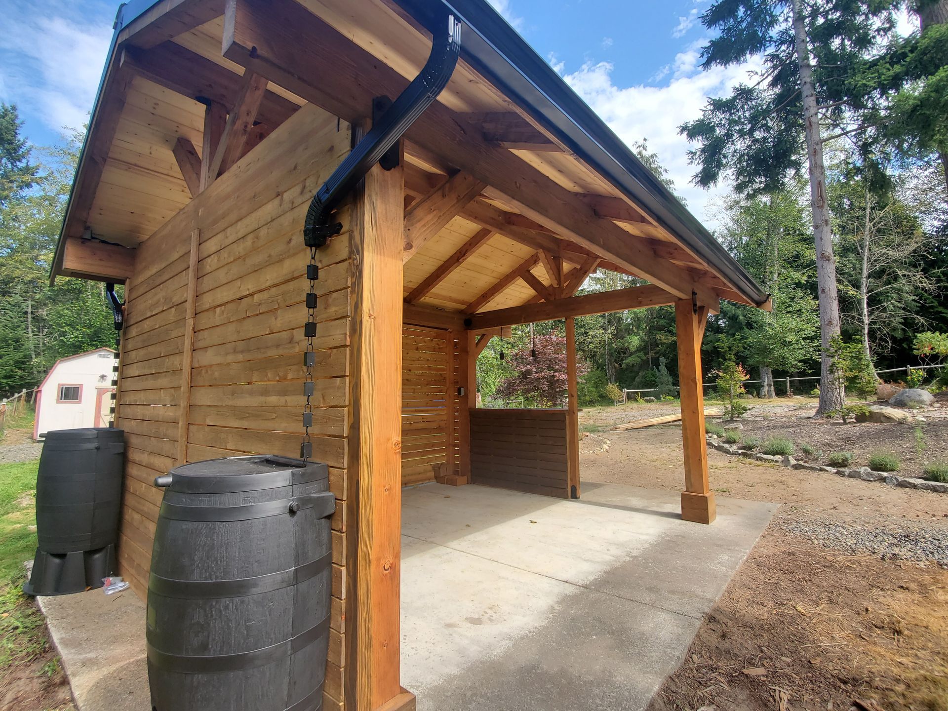 Wooden shed with a covered area, black rain barrel, and gutter in a garden.