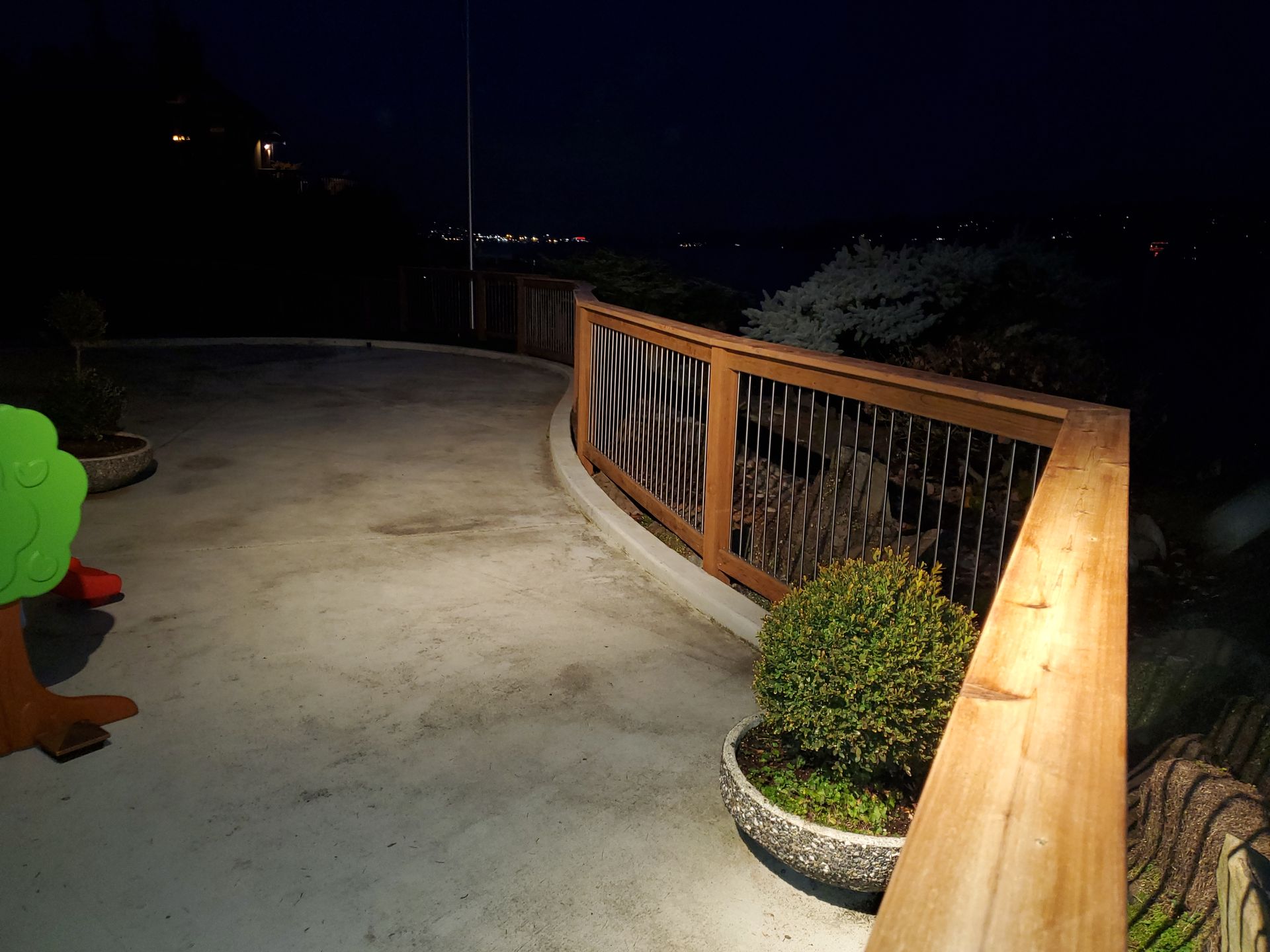 Curved outdoor patio at night, illuminated by soft light. Wooden railing with metal mesh. Small bush in a pot.