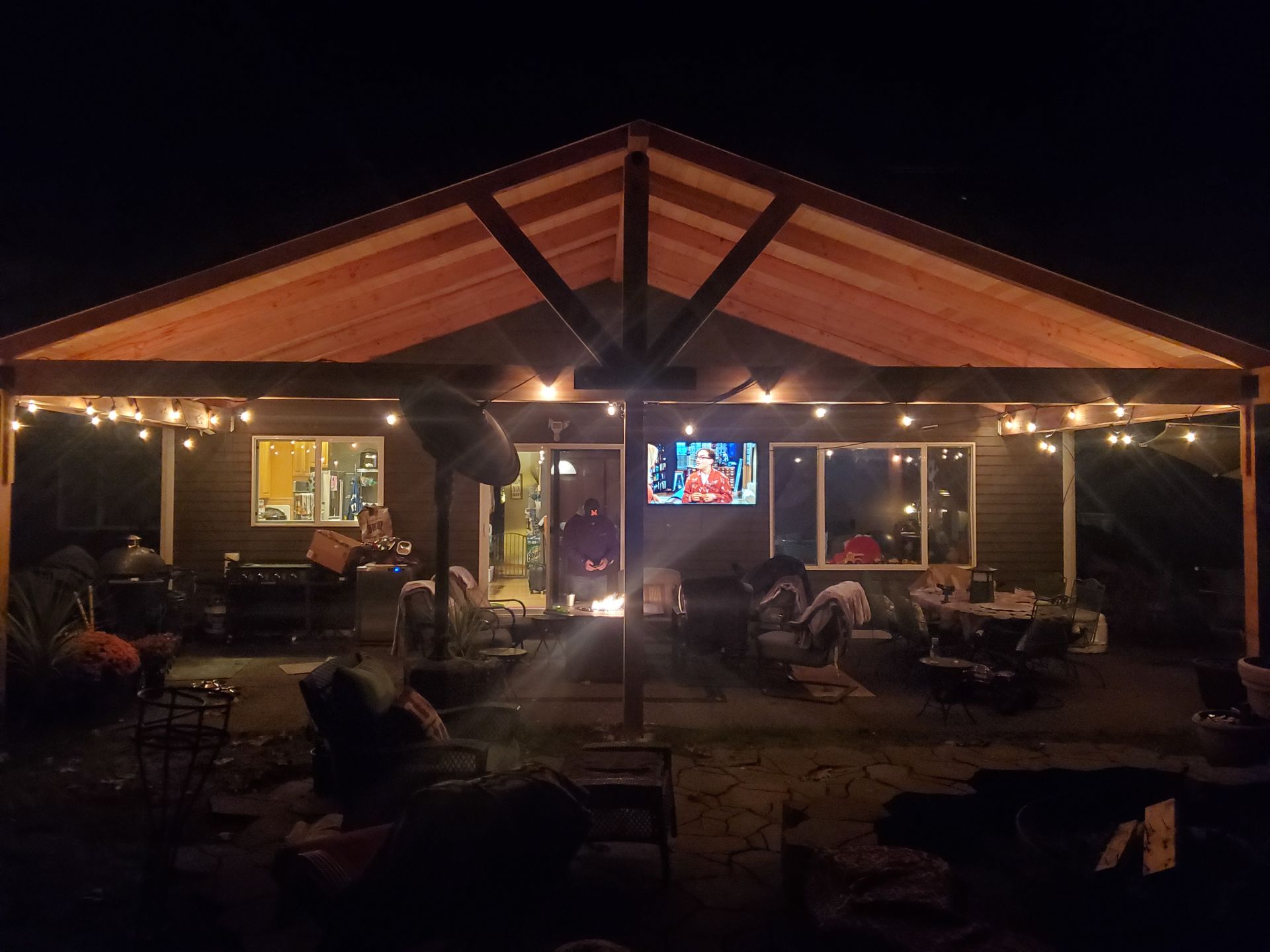 Outdoor patio at night with string lights, TV, and people gathered around a fire.