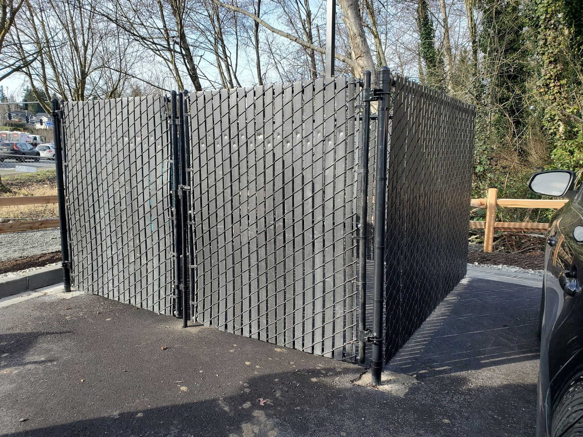 Black chain-link fence with vertical slats encloses a small area, set on asphalt with trees in the background.