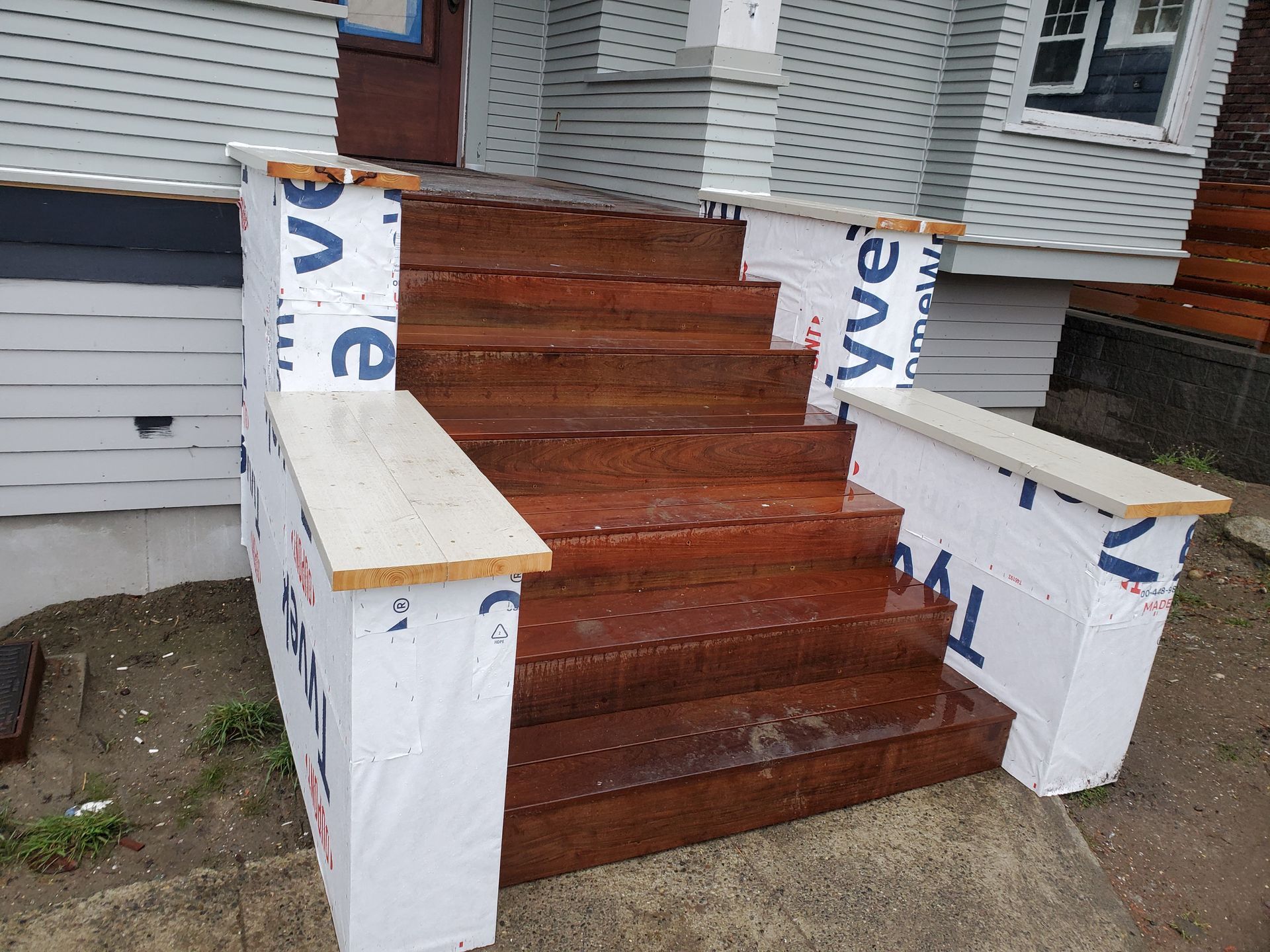 Brown wooden exterior stairs of a house under construction, wrapped in Tyvek with light wood railing caps.