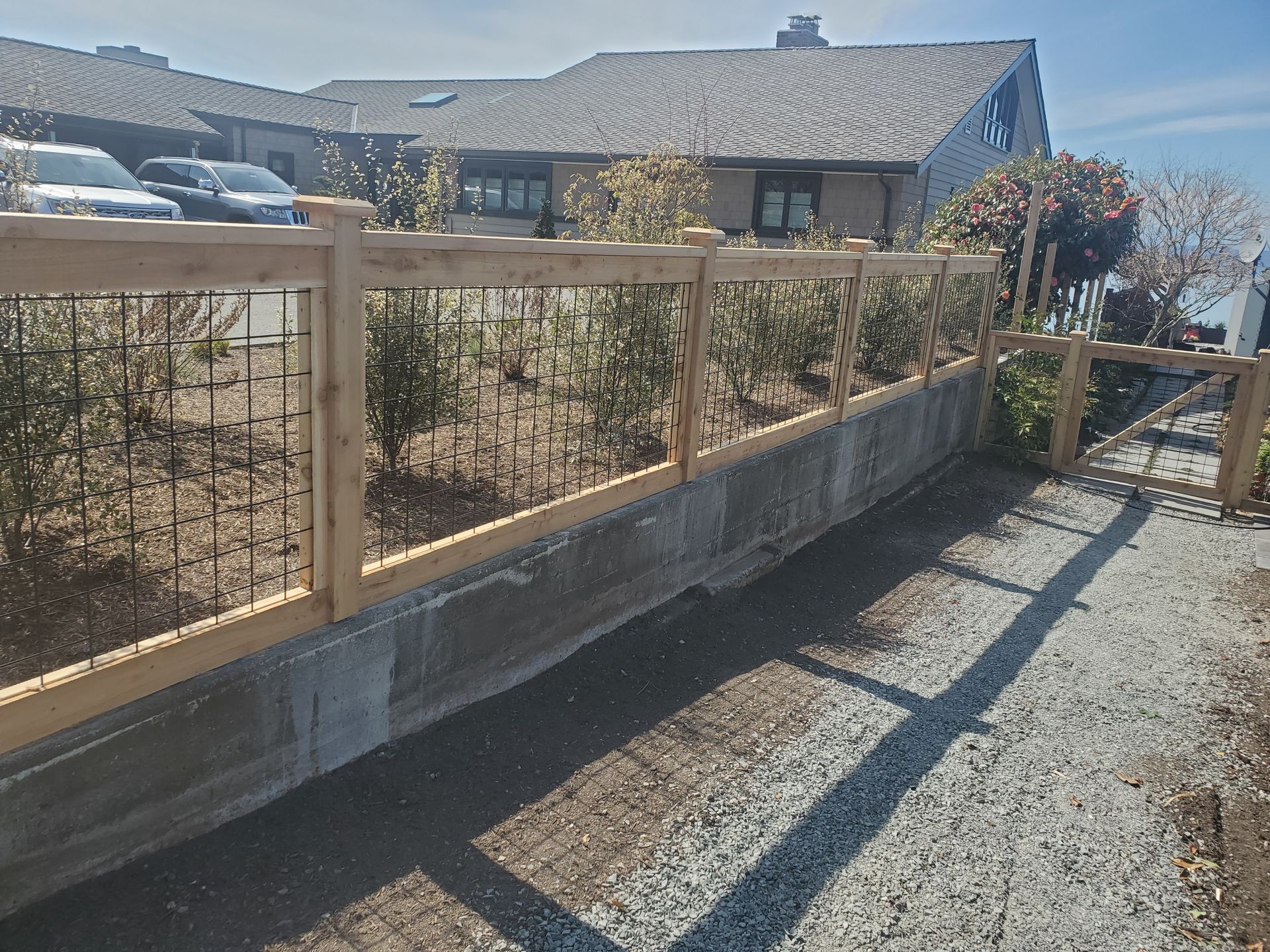 Wooden fence with black metal bars, built on a concrete wall, with plants behind it. Cars and a building in background.