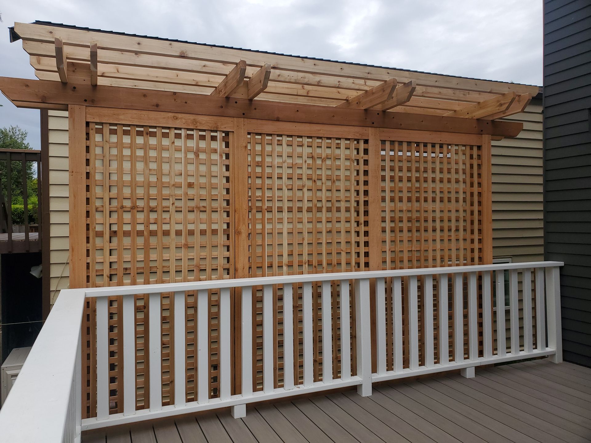 Wooden pergola with lattice panels and white railing on a deck. Brown siding in the background.
