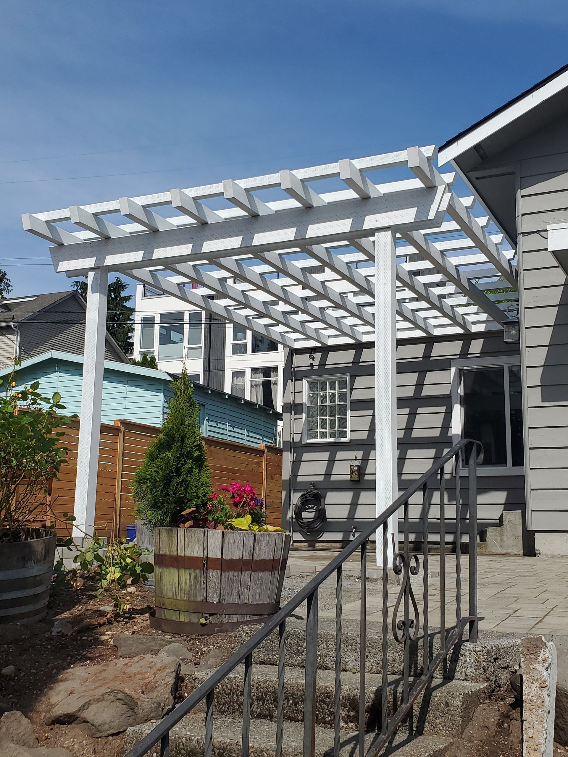 White pergola attached to a gray house, over a patio with barrel planters and a wrought iron railing.
