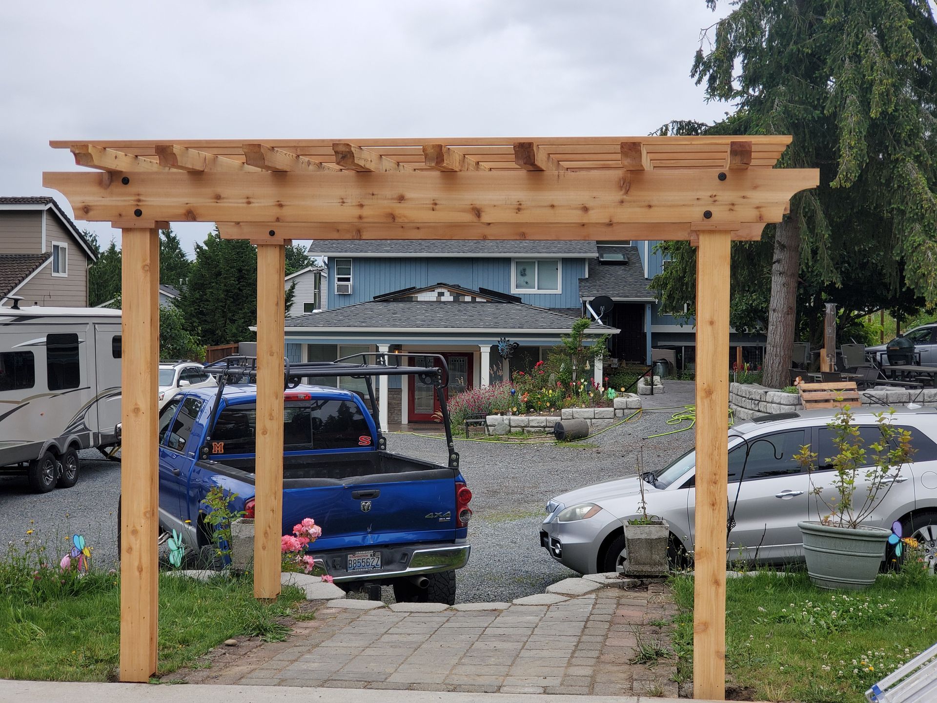 Wooden pergola framing a paved pathway leading towards a house with vehicles parked nearby.