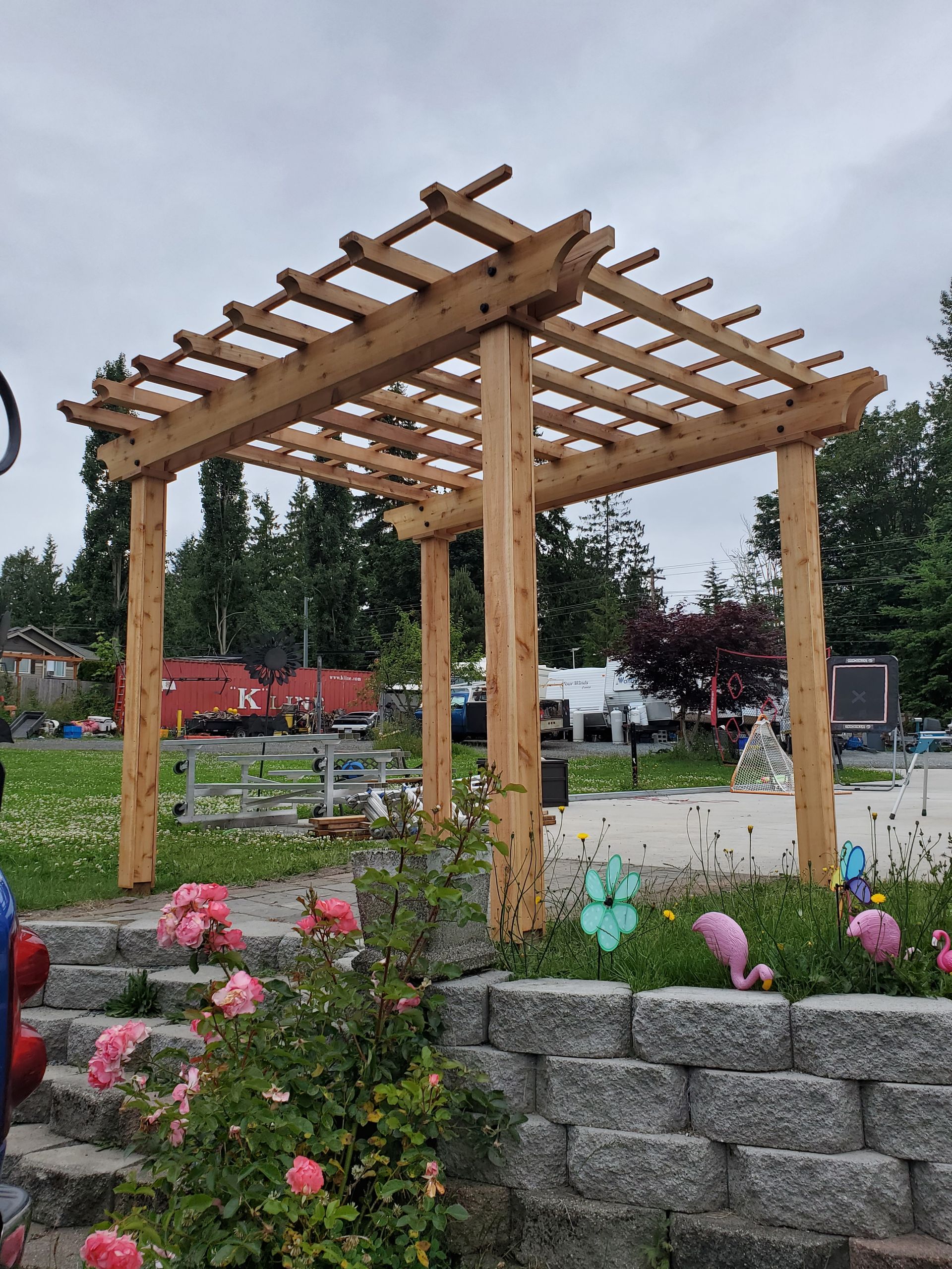 Wooden pergola with latticed roof in a garden, with flowers and a stone retaining wall.