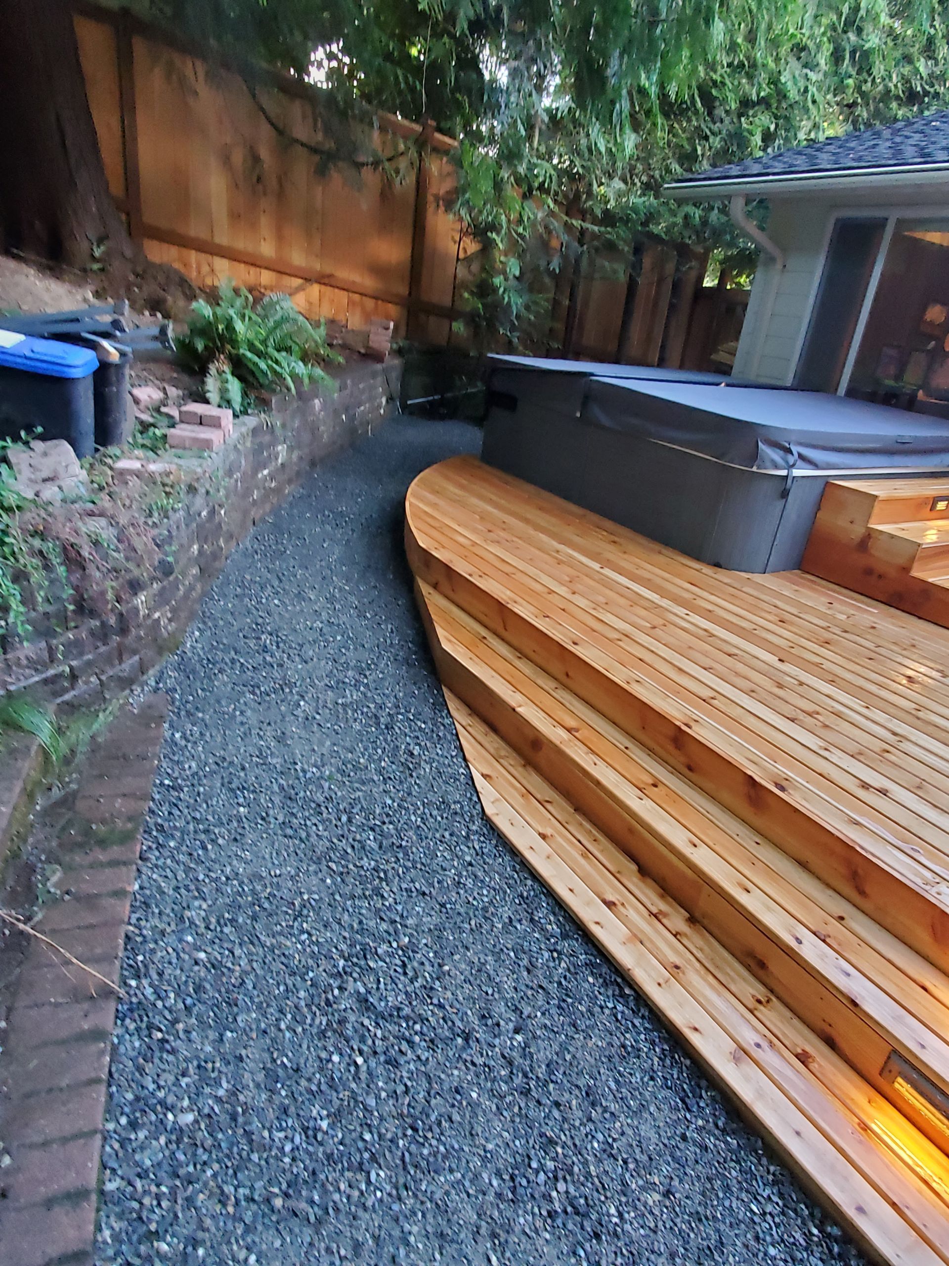 Gravel path next to a wooden deck with steps, a hot tub, and a retaining wall.