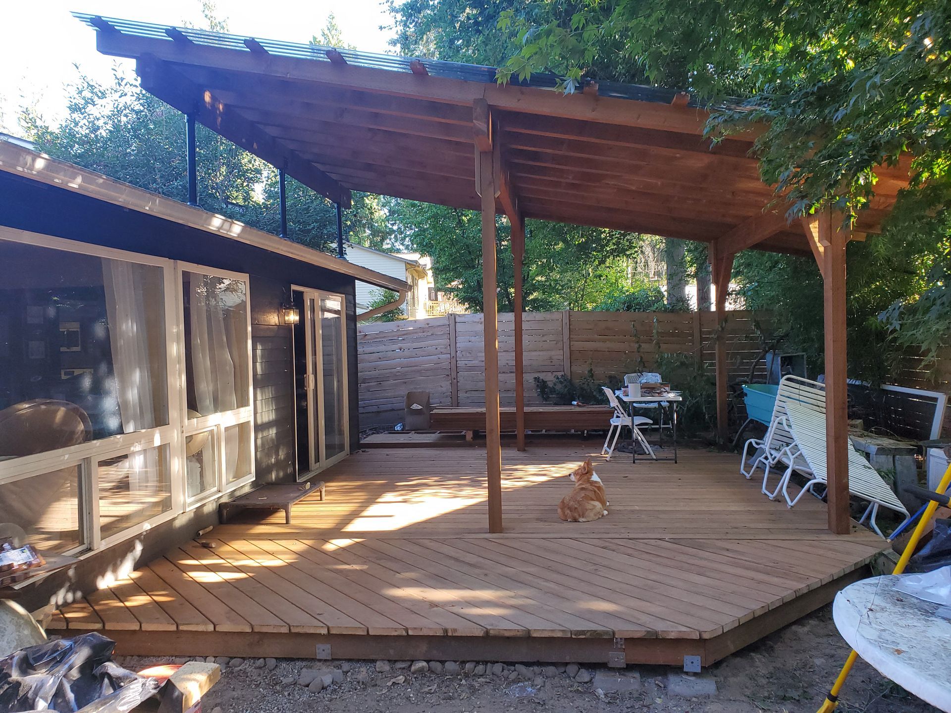 Wooden deck with attached roof, dog sits in the center. House on left, fence and trees in background.