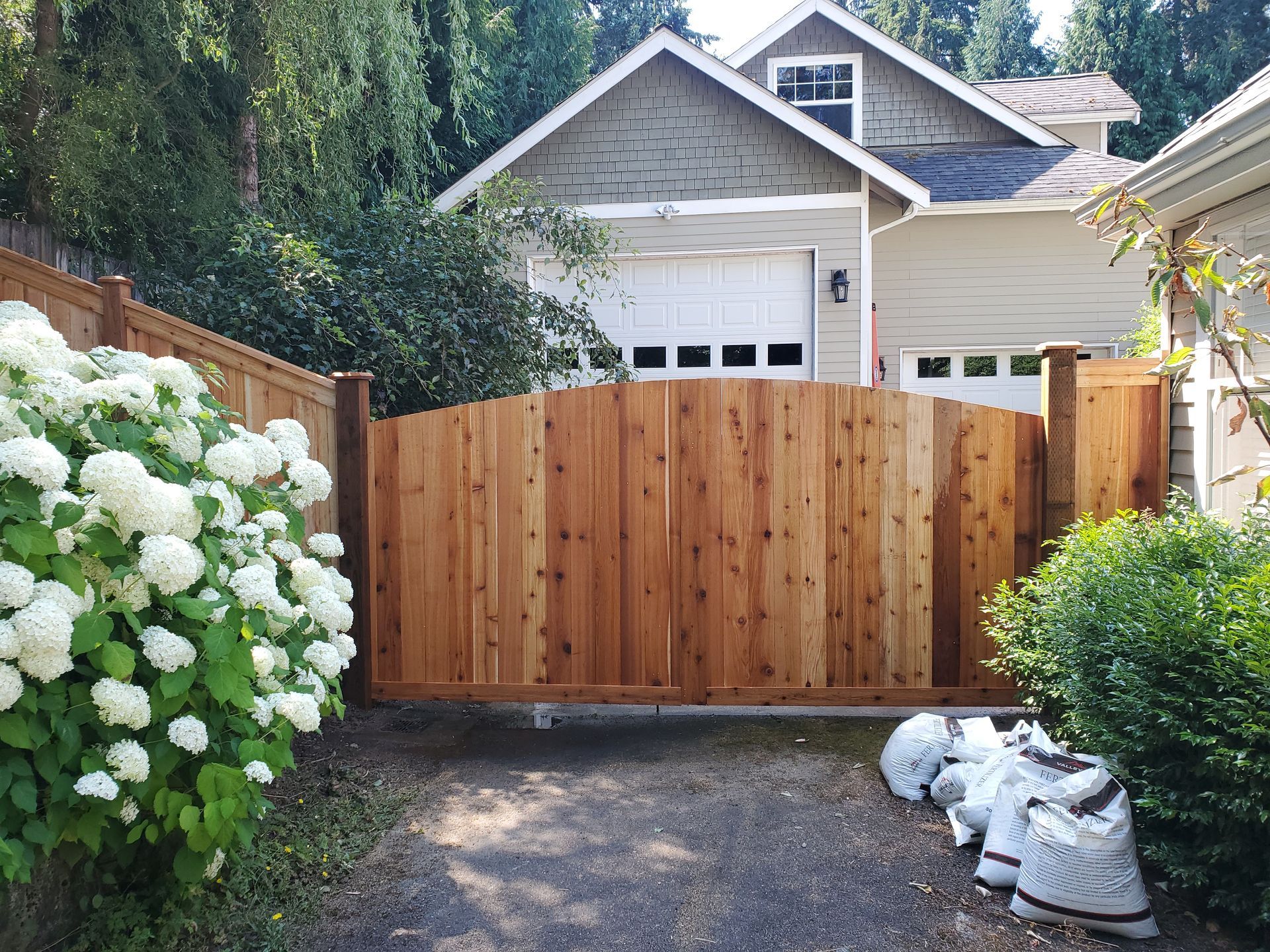 Wooden arched gate in front of a house, with hydrangeas on the left, driveway, and bags on the right.