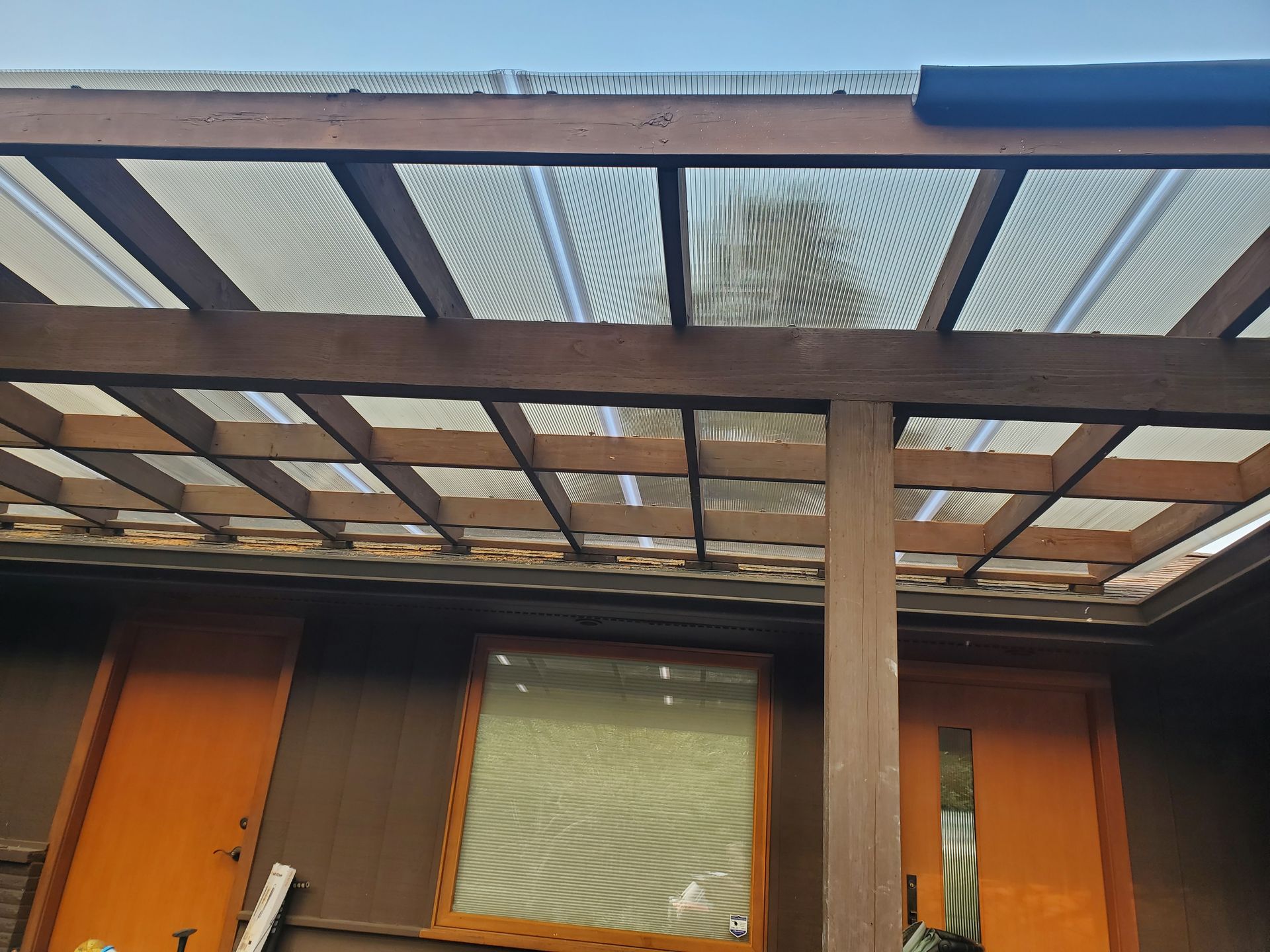 Wooden pergola with clear roofing over a brown building with orange doors and a window.