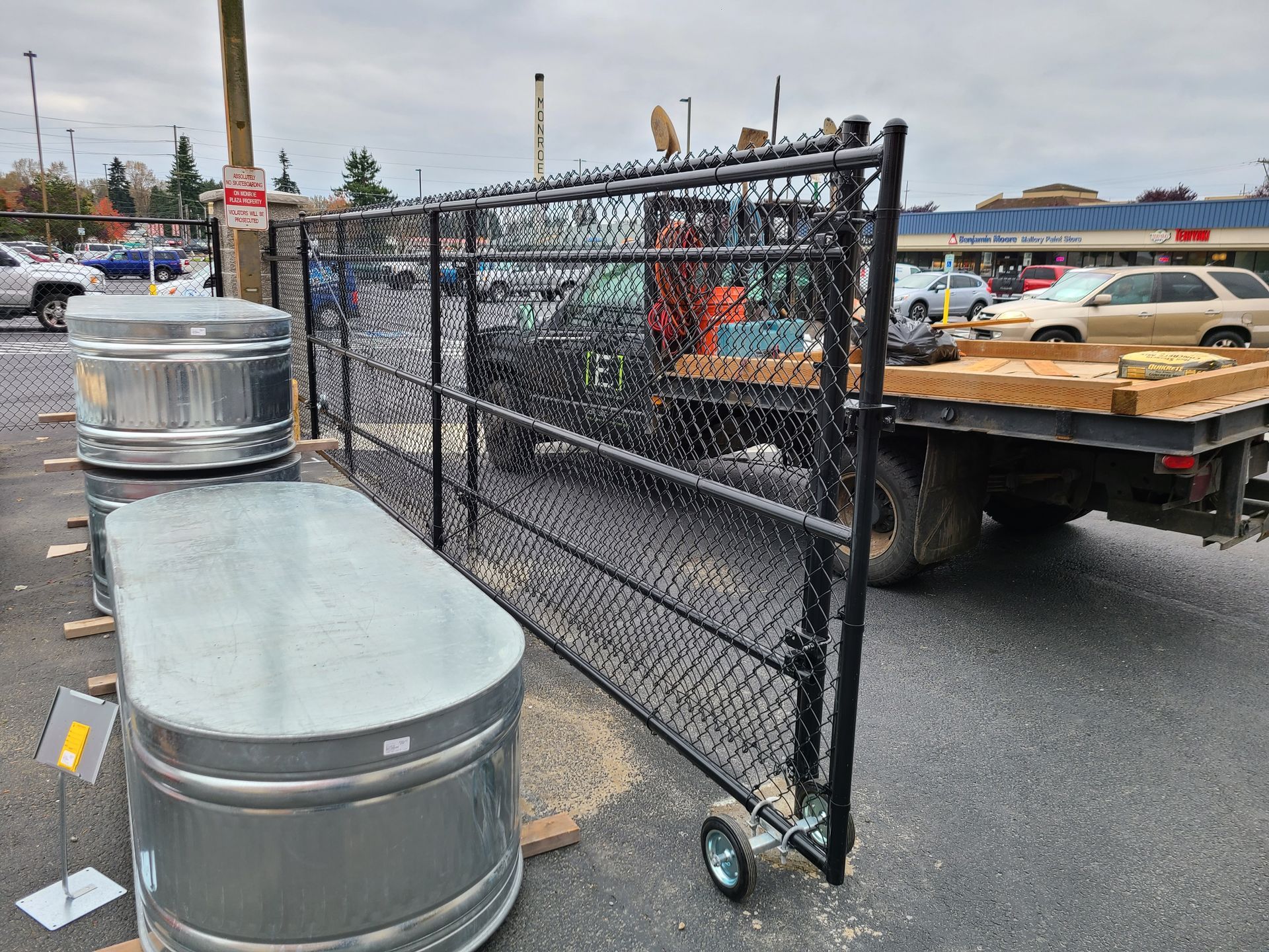Black chain-link fence on wheels next to metal water tanks, parked outside; cars and buildings in the background.